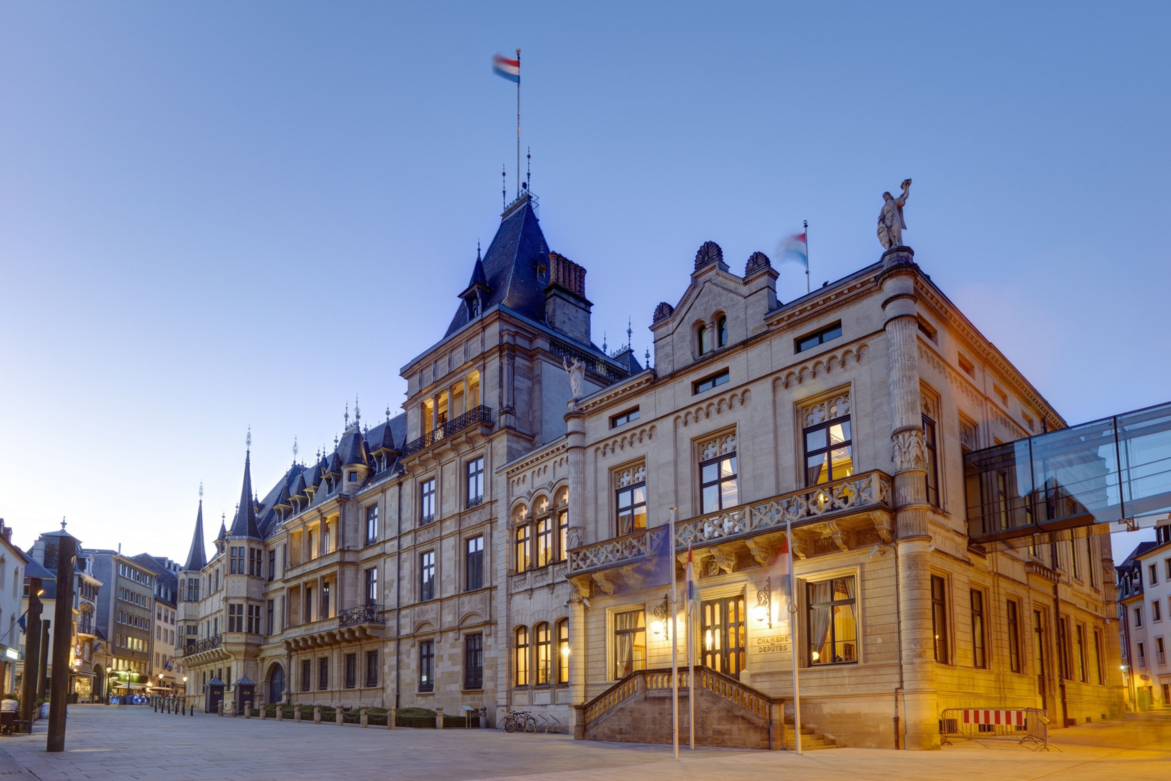 The image shows a grand historic building with ornate architectural details, peaked roofs, and flags. It's illuminated against a twilight sky, creating a majestic scene.