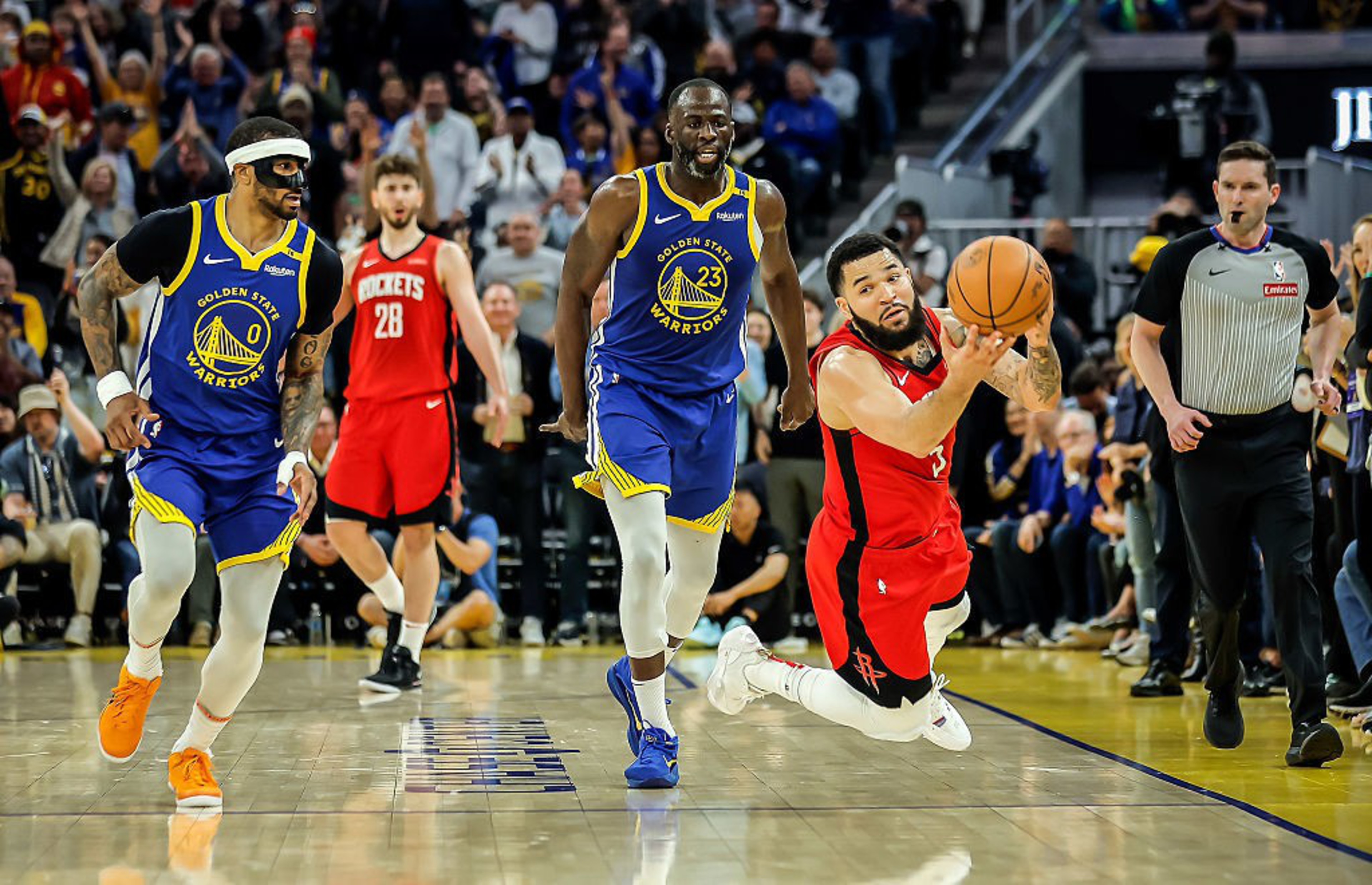 A basketball player in a red jersey dives to reach a ball while two players in blue jerseys run behind him. An official watches the action closely.