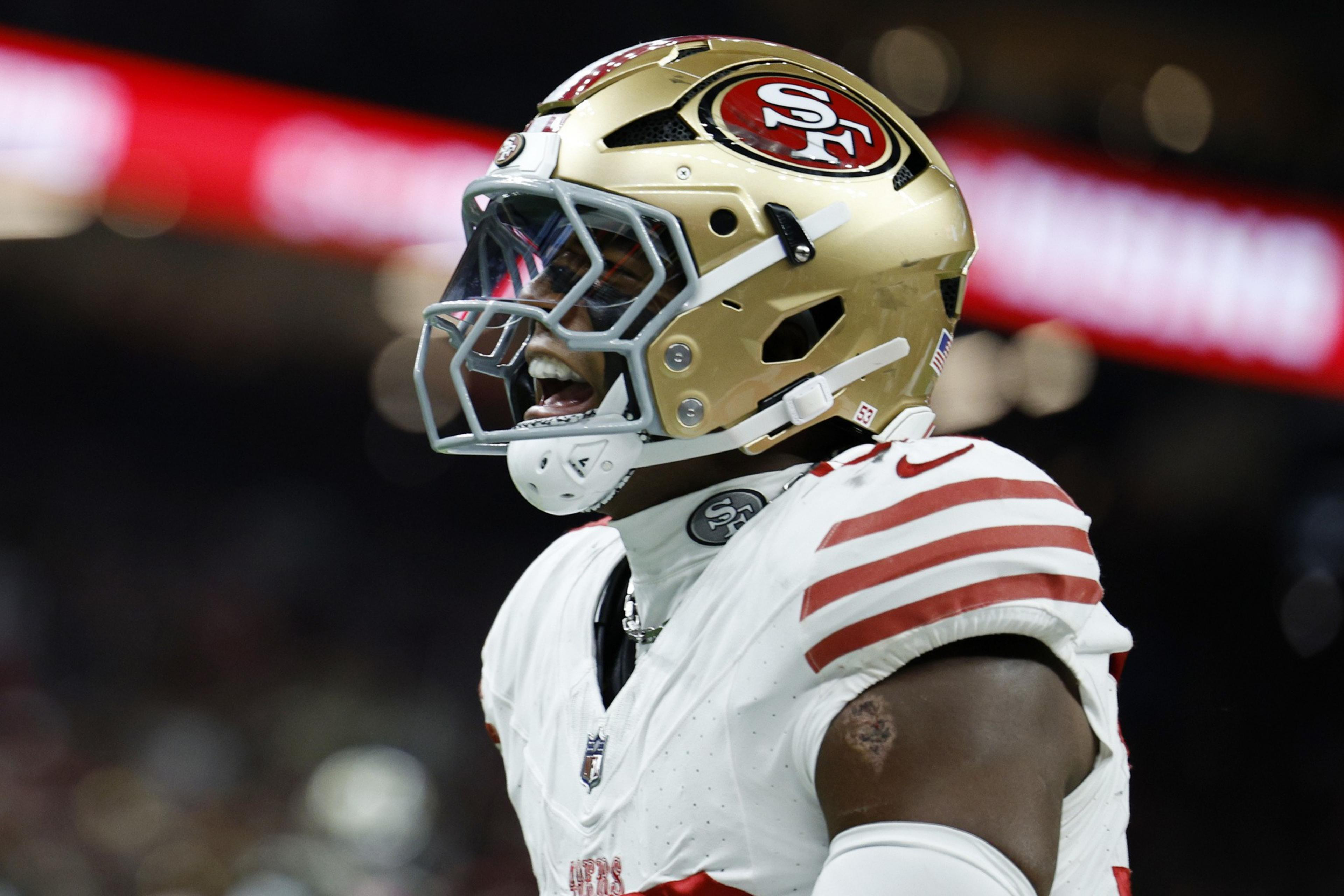 A San Francisco 49ers football player in full uniform and helmet celebrates on the field with his mouth open.