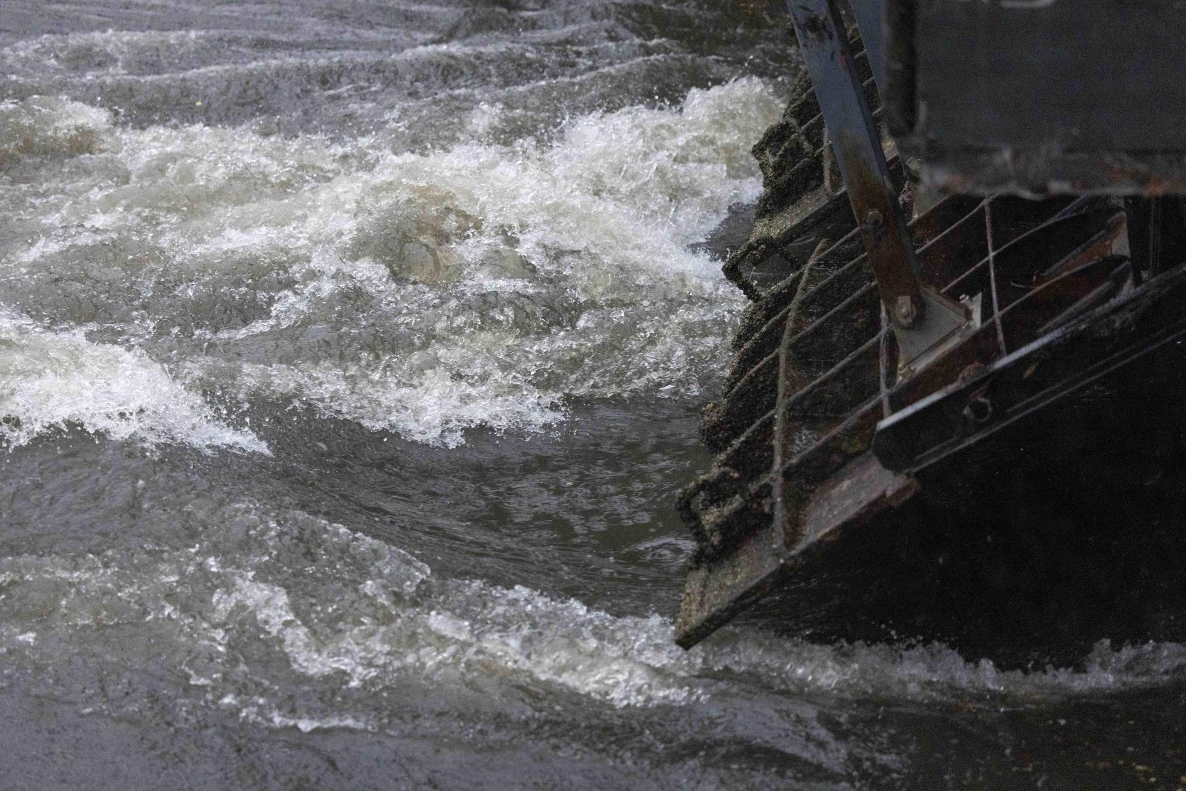 The image shows a turbulent river with foamy white water swirling around a partially submerged metal waterwheel.
