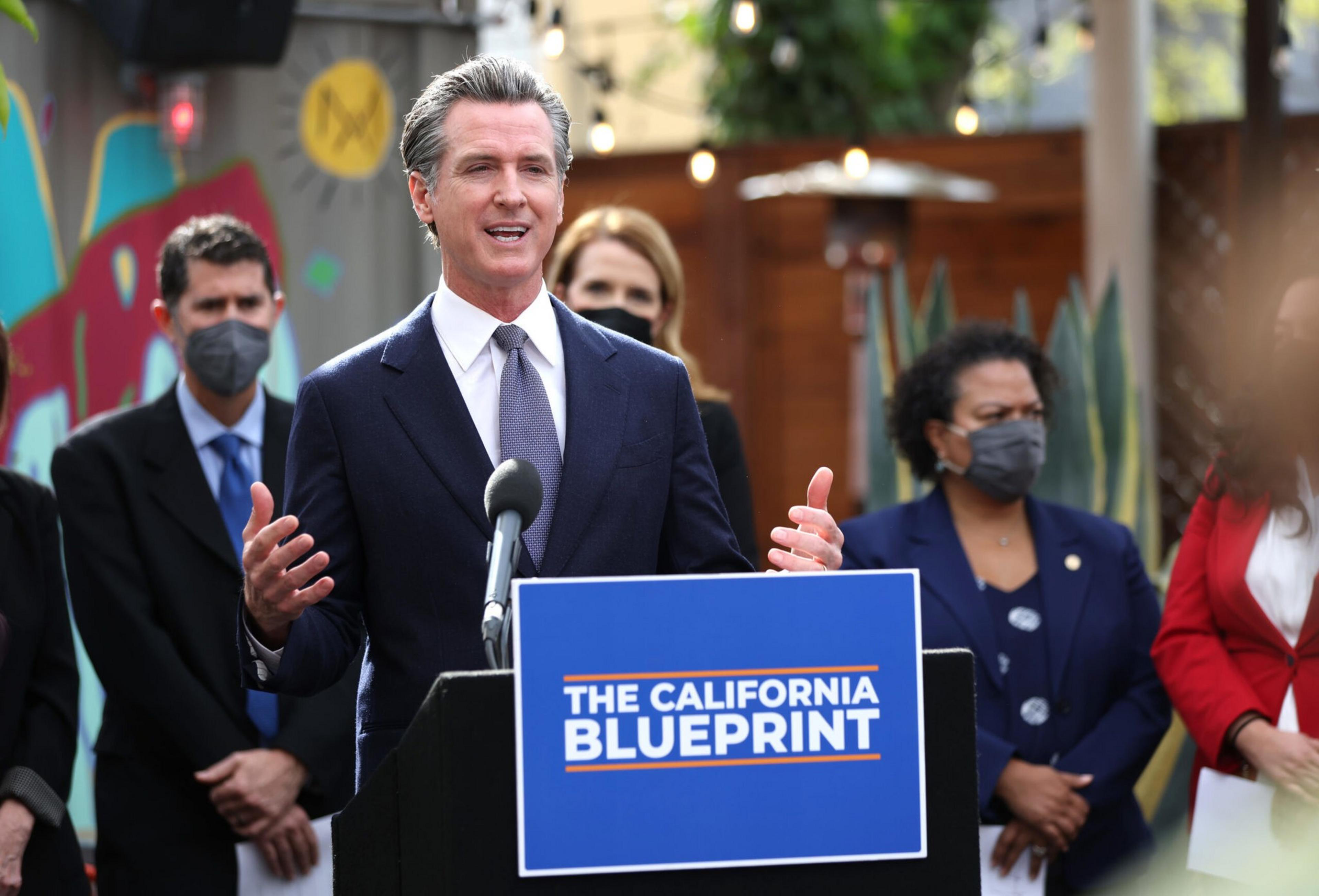 A man in a suit speaks at a podium labeled "THE CALIFORNIA BLUEPRINT" with four masked people standing behind him.