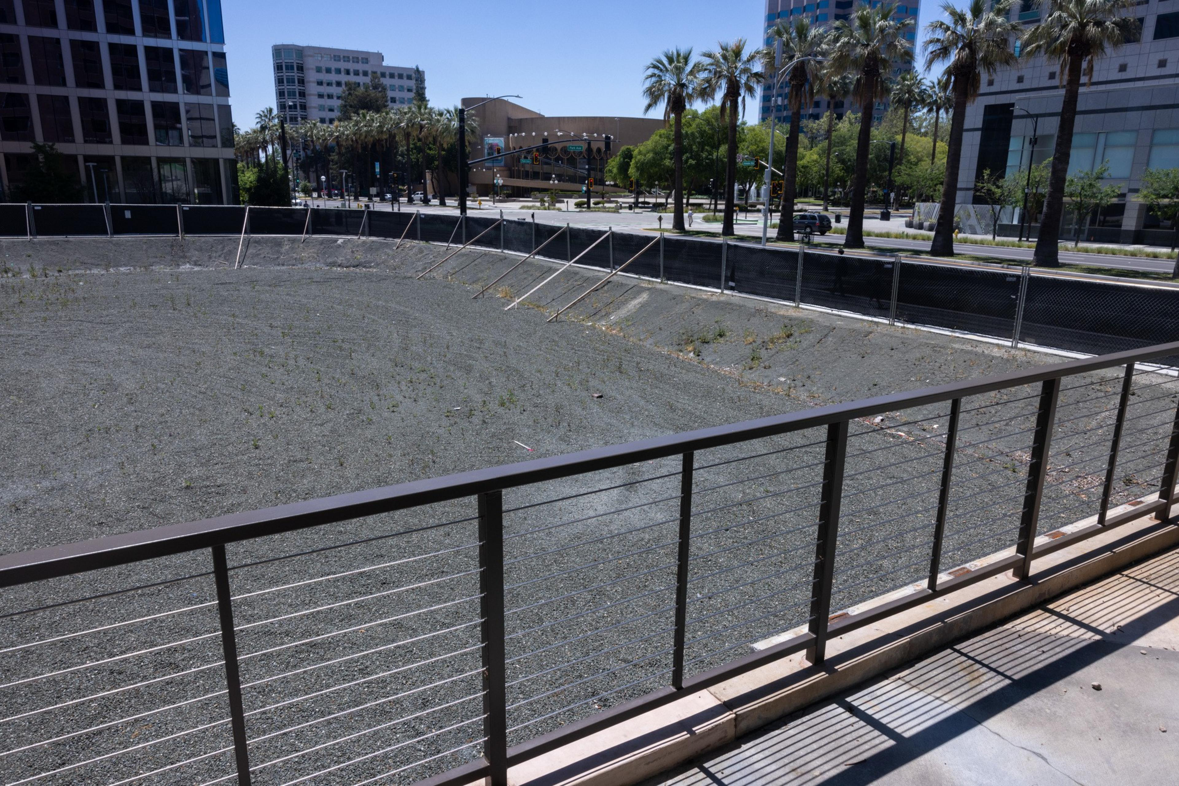 The image shows a fenced-off, rectangular, gravel-filled excavation site surrounded by modern buildings and palm trees under a clear blue sky.