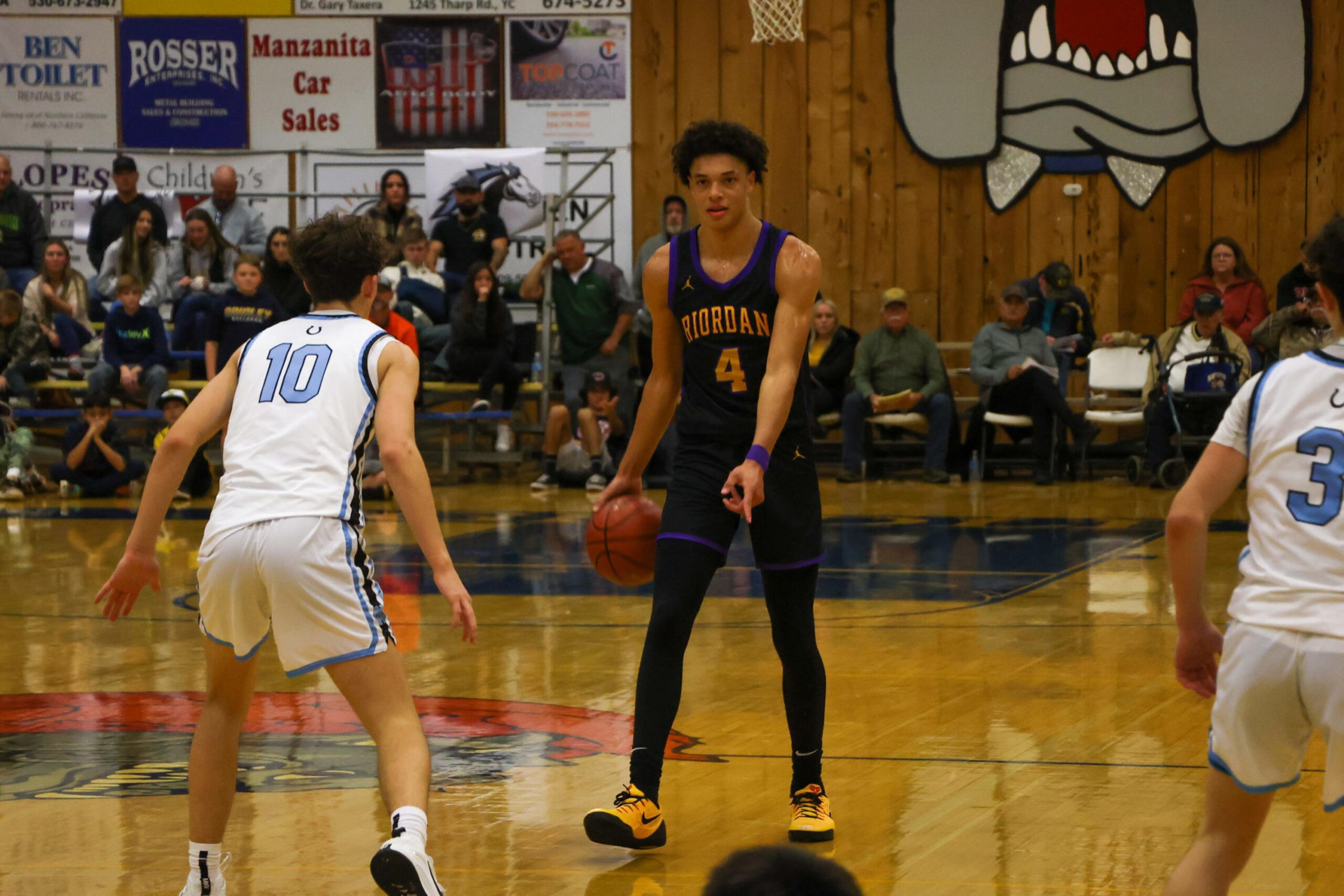 A basketball player in a black uniform with "Riordan" and number 4 dribbles the ball while two players in white defend on a gym court.