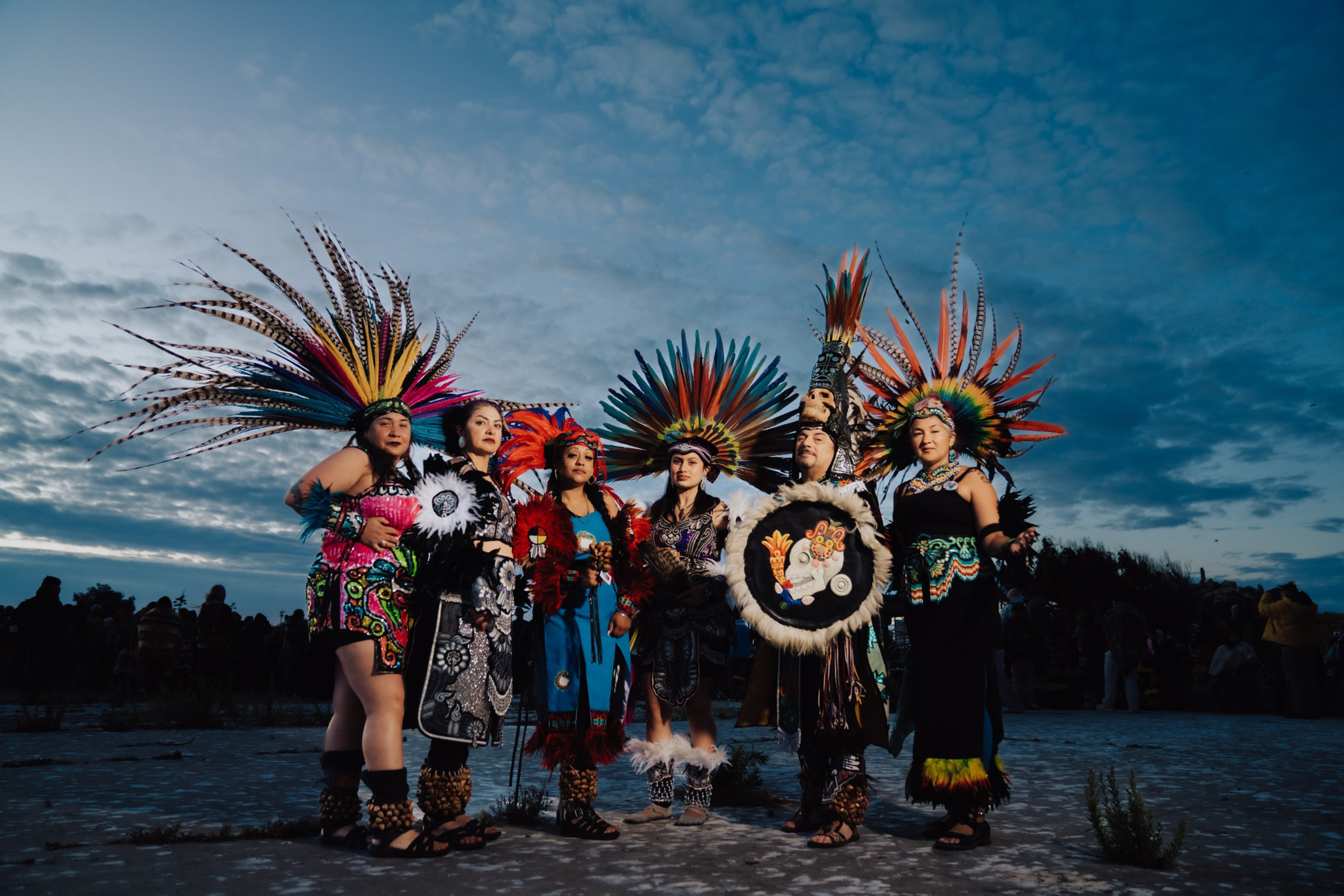 Six people stand together outdoors at dusk, wearing traditional colorful Aztec feathered headdresses and vibrant ceremonial clothing.