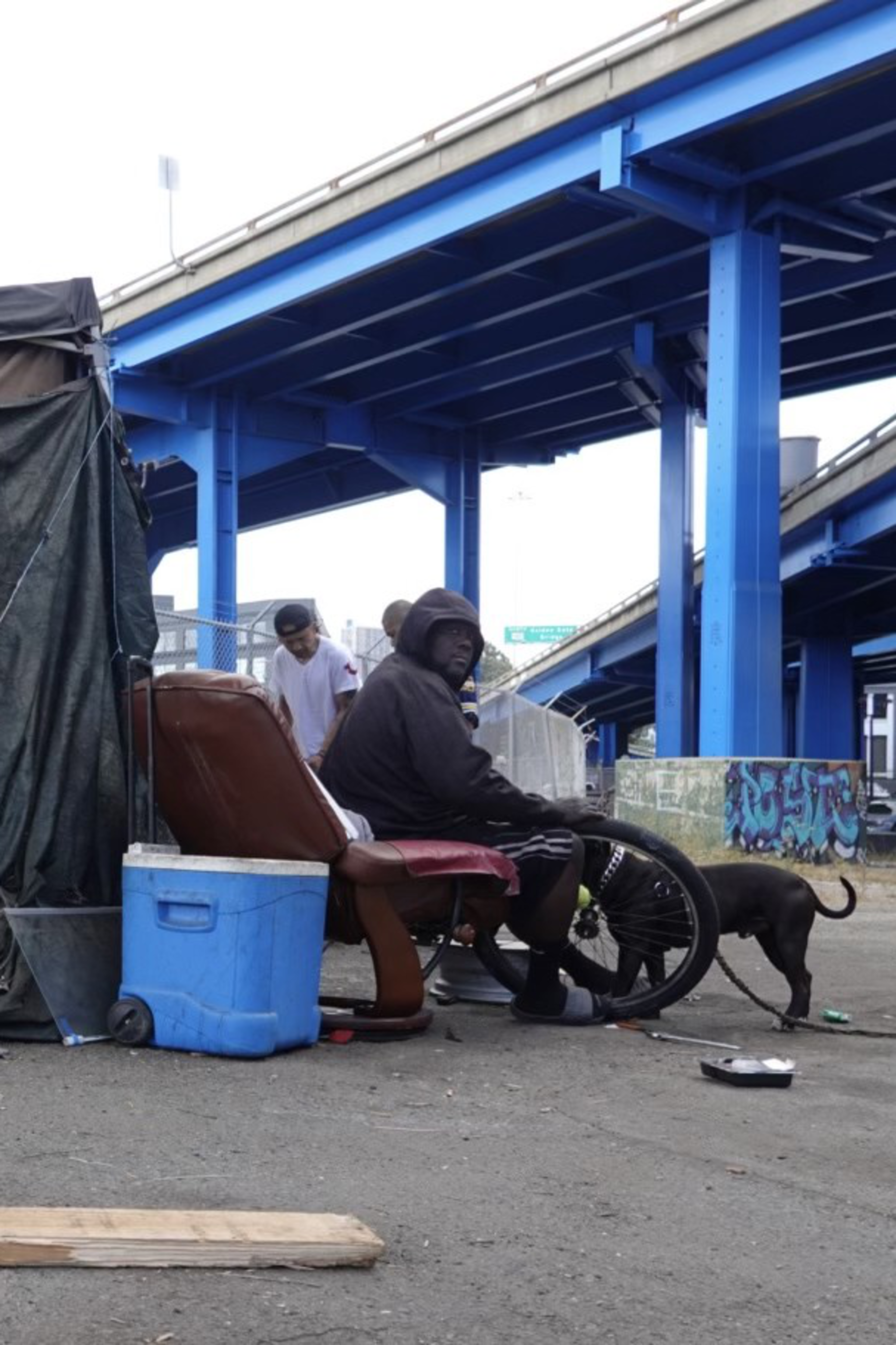 A man sits on a recliner under a blue overpass, next to makeshift shelter materials and a blue cooler. Another person and a dog are nearby on the concrete ground.