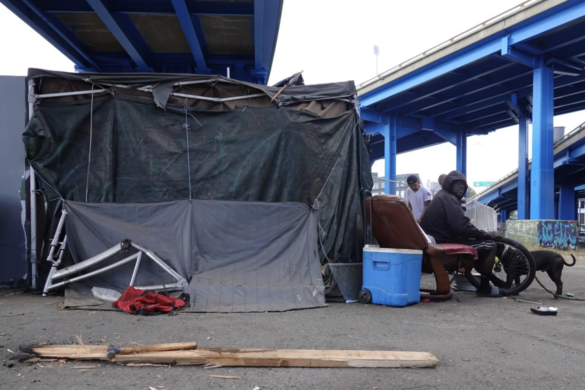 A man sits on a recliner under a blue overpass, next to makeshift shelter materials and a blue cooler. Another person and a dog are nearby on the concrete ground.