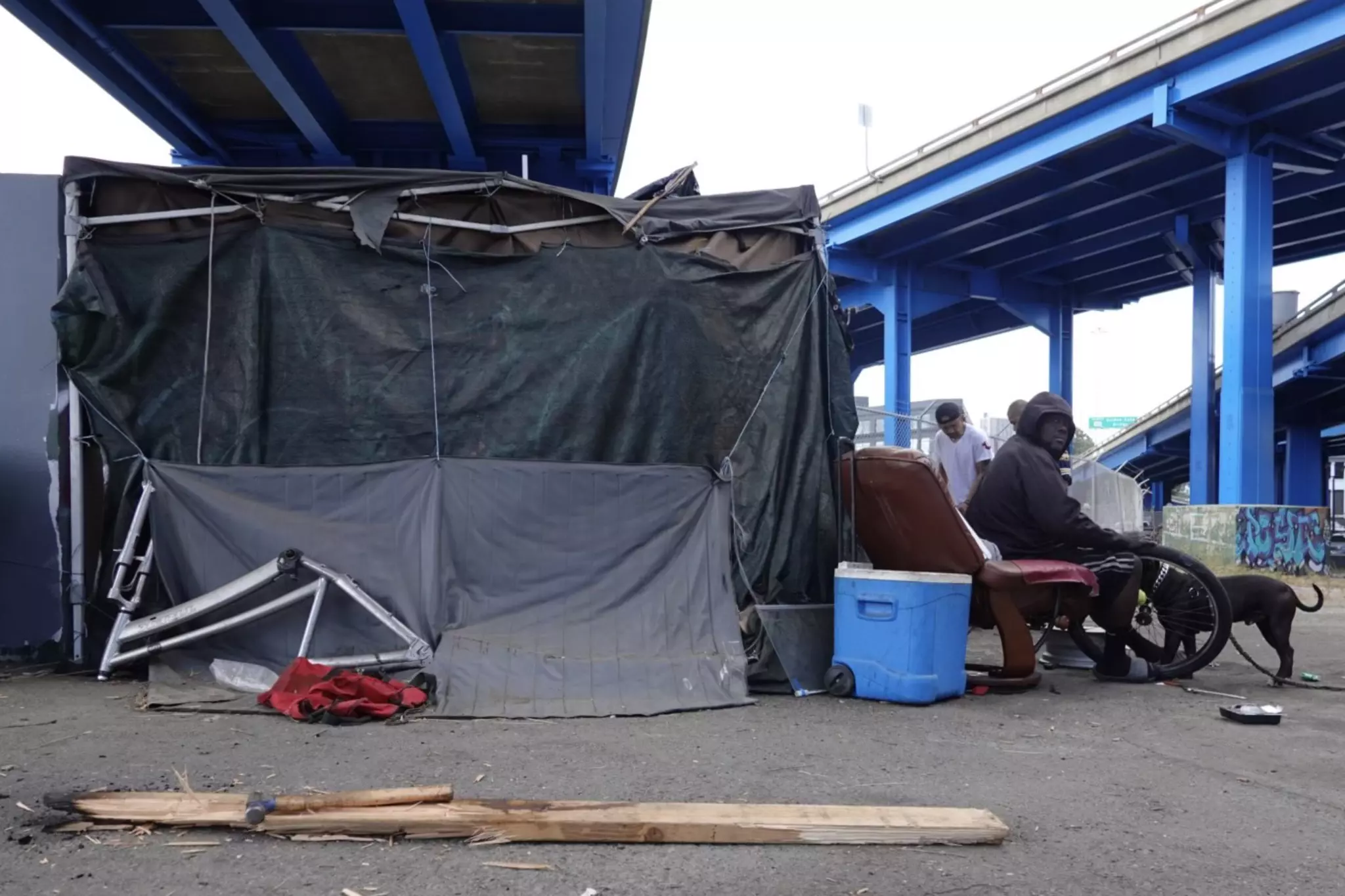 A man sits on a recliner under a blue overpass, next to makeshift shelter materials and a blue cooler. Another person and a dog are nearby on the concrete ground.
