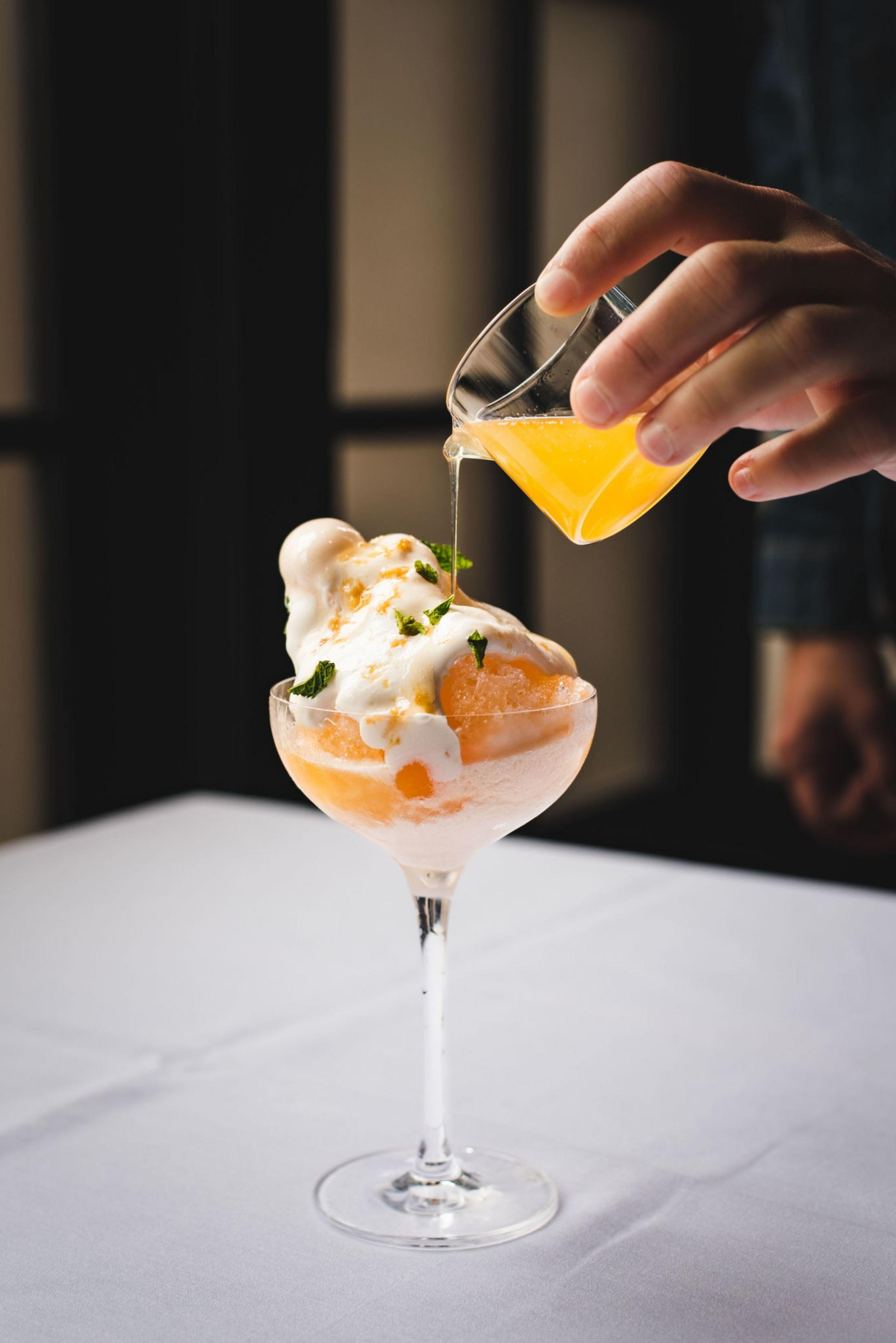 An elegant shave ice dessert in a coupe. A hand pours syrup over the top.