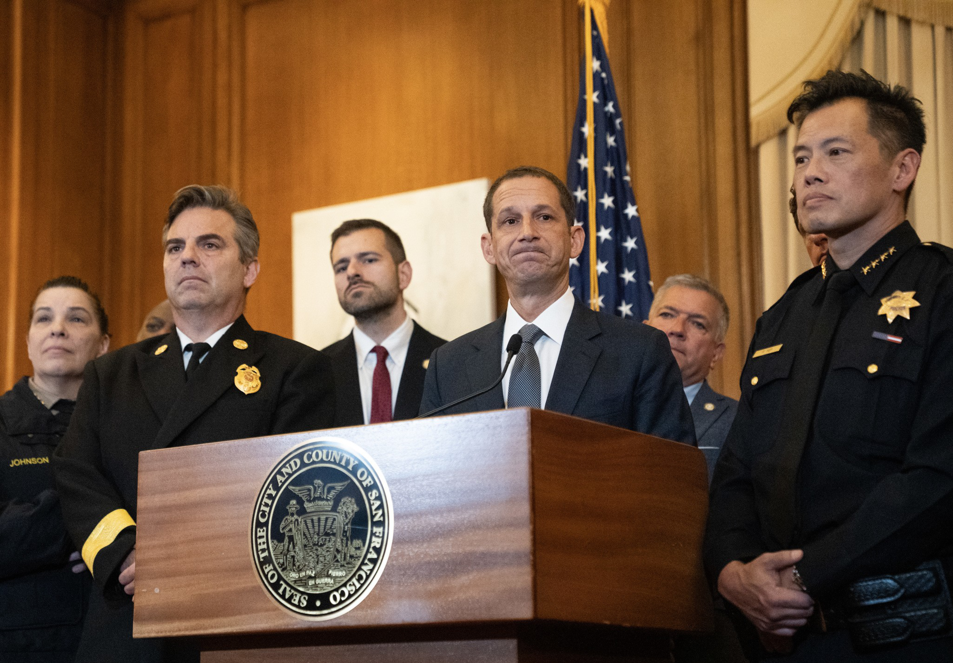 A group of officials, including firefighters, police officers, and a suited man, stand behind a podium with a San Francisco city seal and US flag.