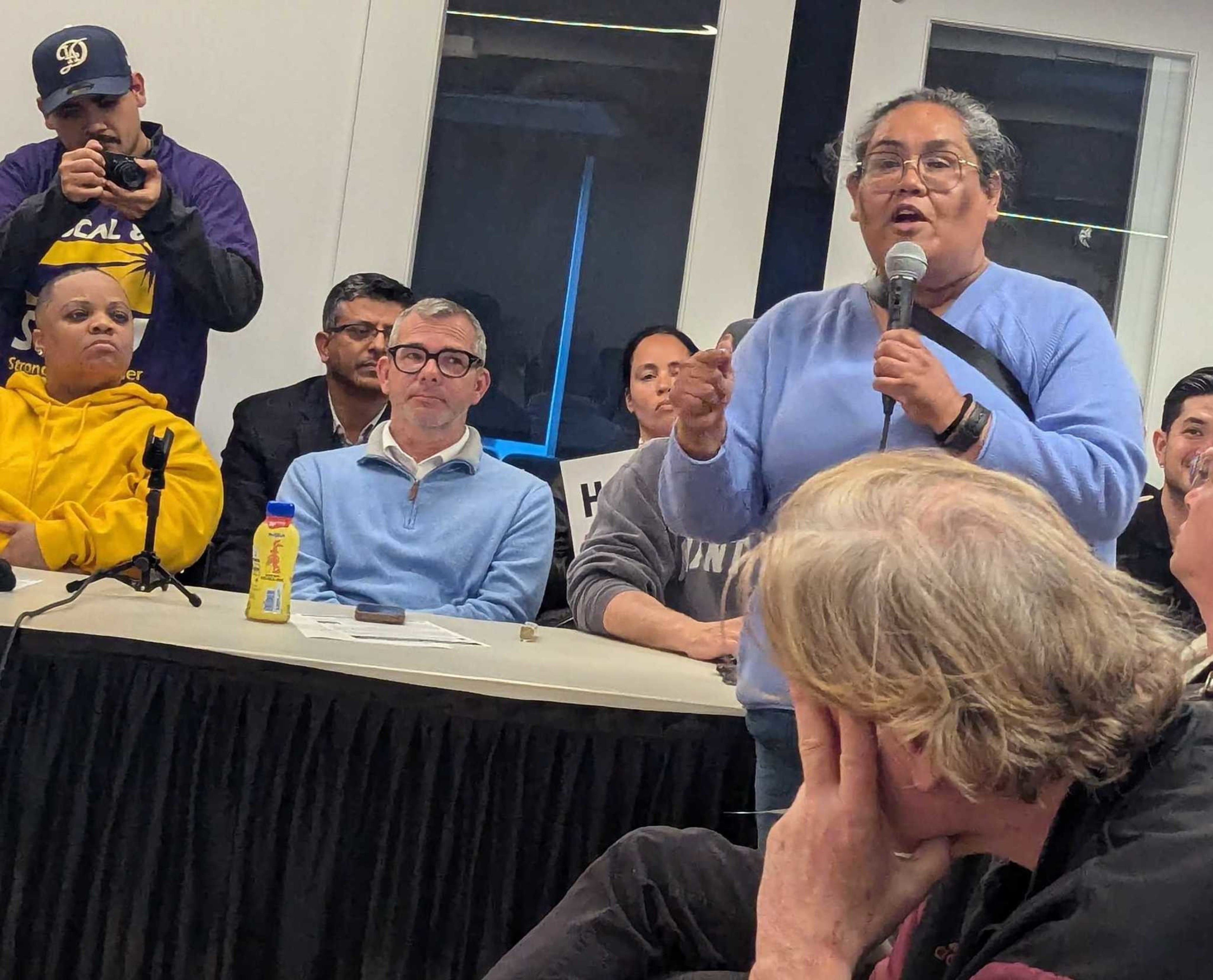 A woman in a blue sweater speaks at a microphone as people seated around her listen.