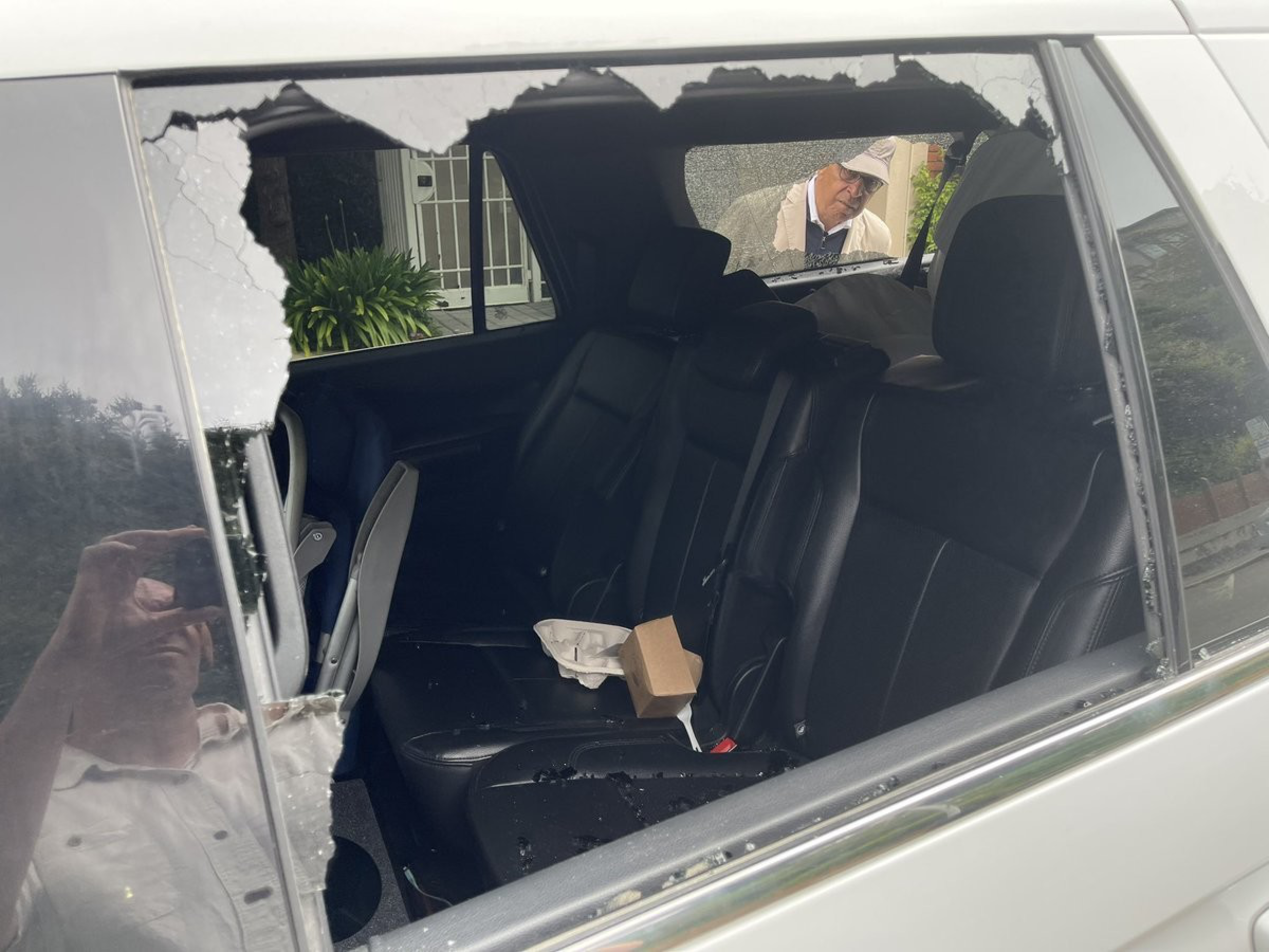 A car’s rear passenger window is shattered, with broken glass on the black seats inside, where a small box and paper container rest; a man peers inside from outside.