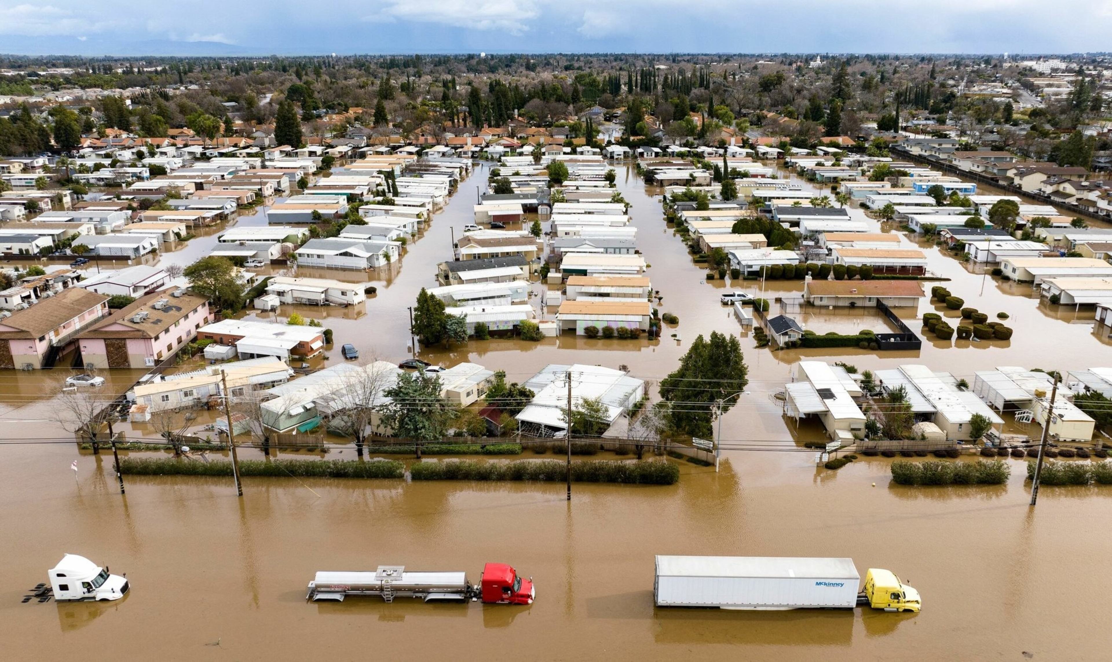 A residential area is flooded with murky brown water, submerging streets and the lower parts of houses, while three trucks partially drive through the flood.