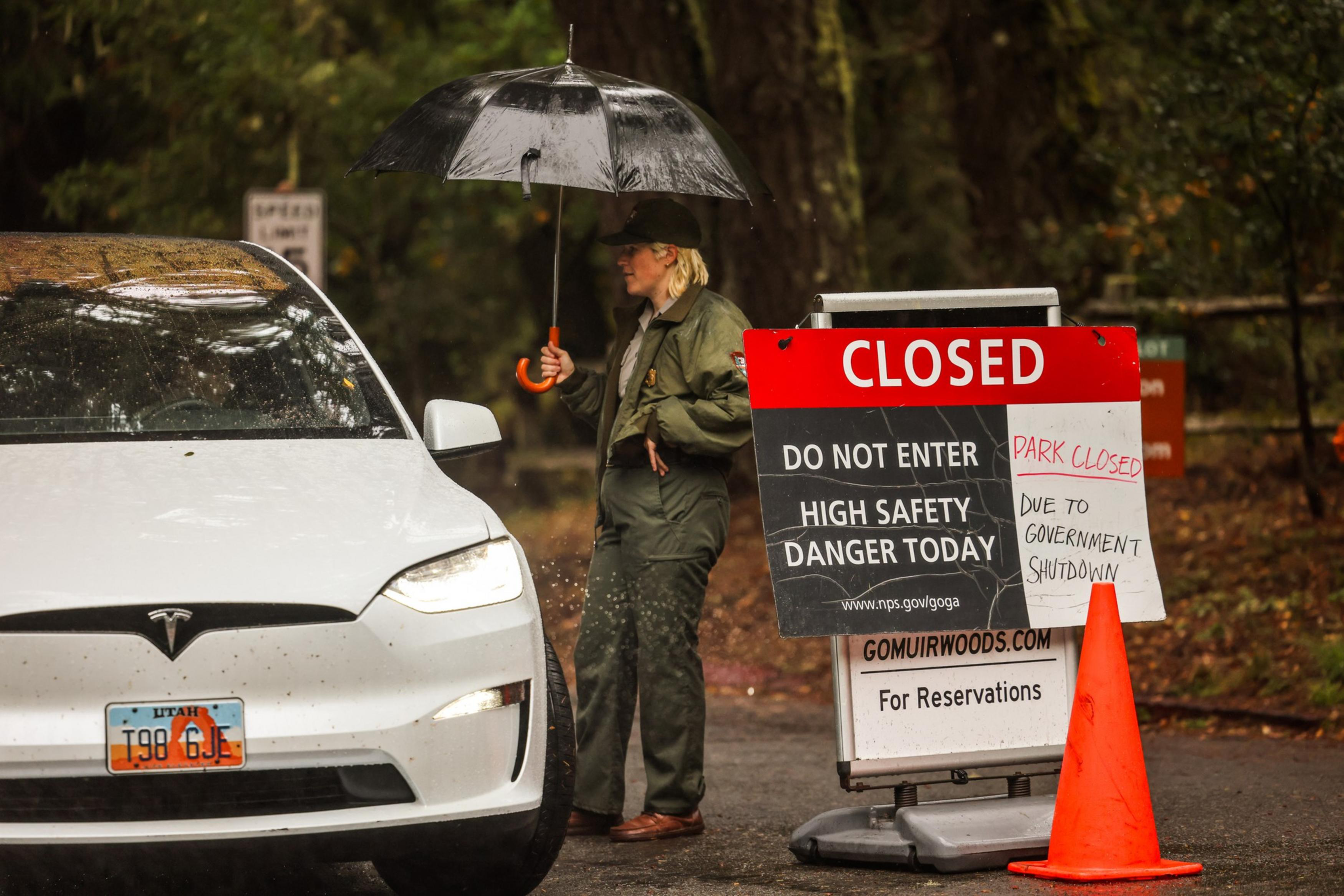 A woman holds a black umbrella while talking to the driver of a white Tesla near a red and black “CLOSED” sign warning of high safety danger and government shutdown.