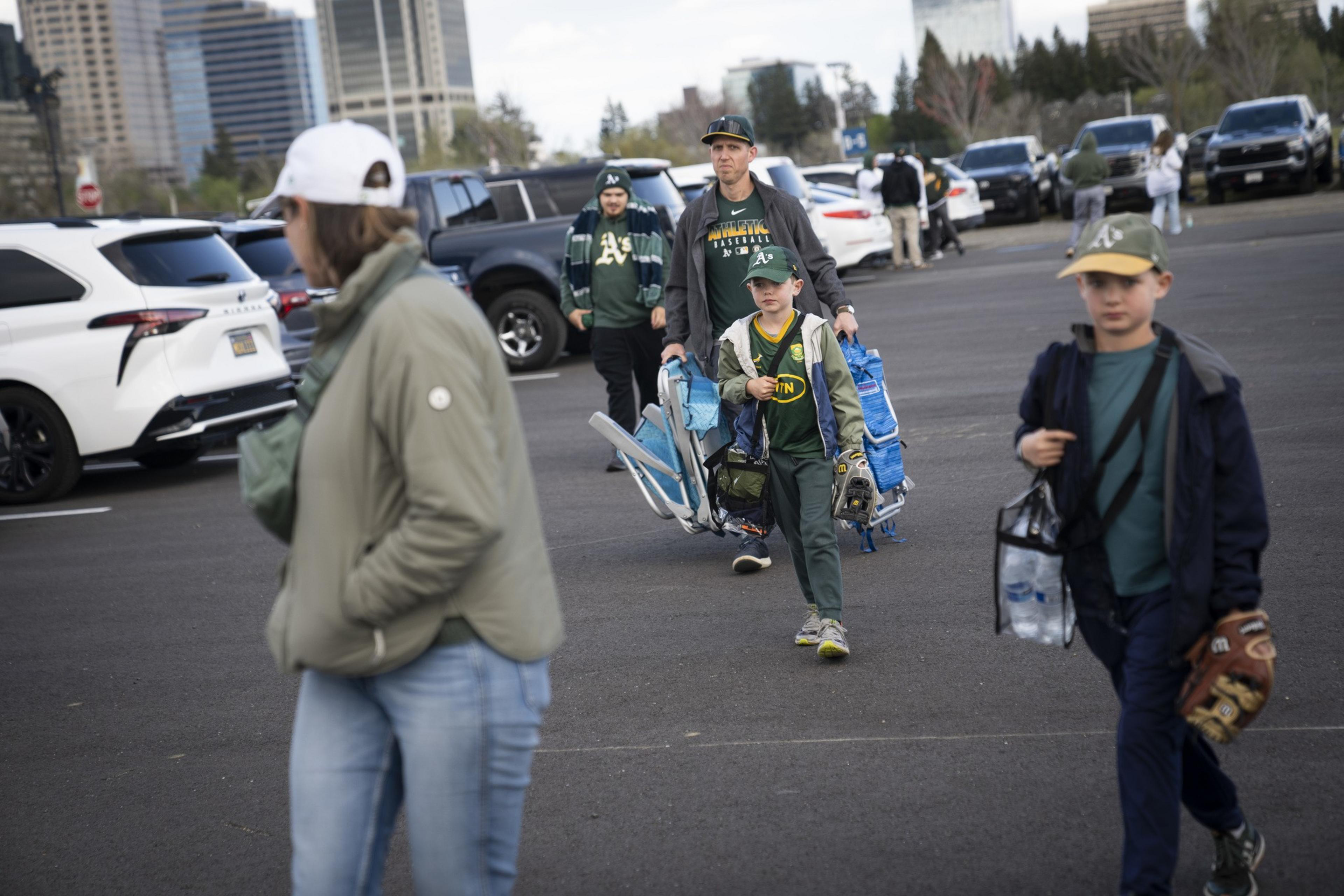 Several people walk through a parking lot carrying baseball gear. They are wearing green and yellow apparel. Cars and a city skyline are in the background.
