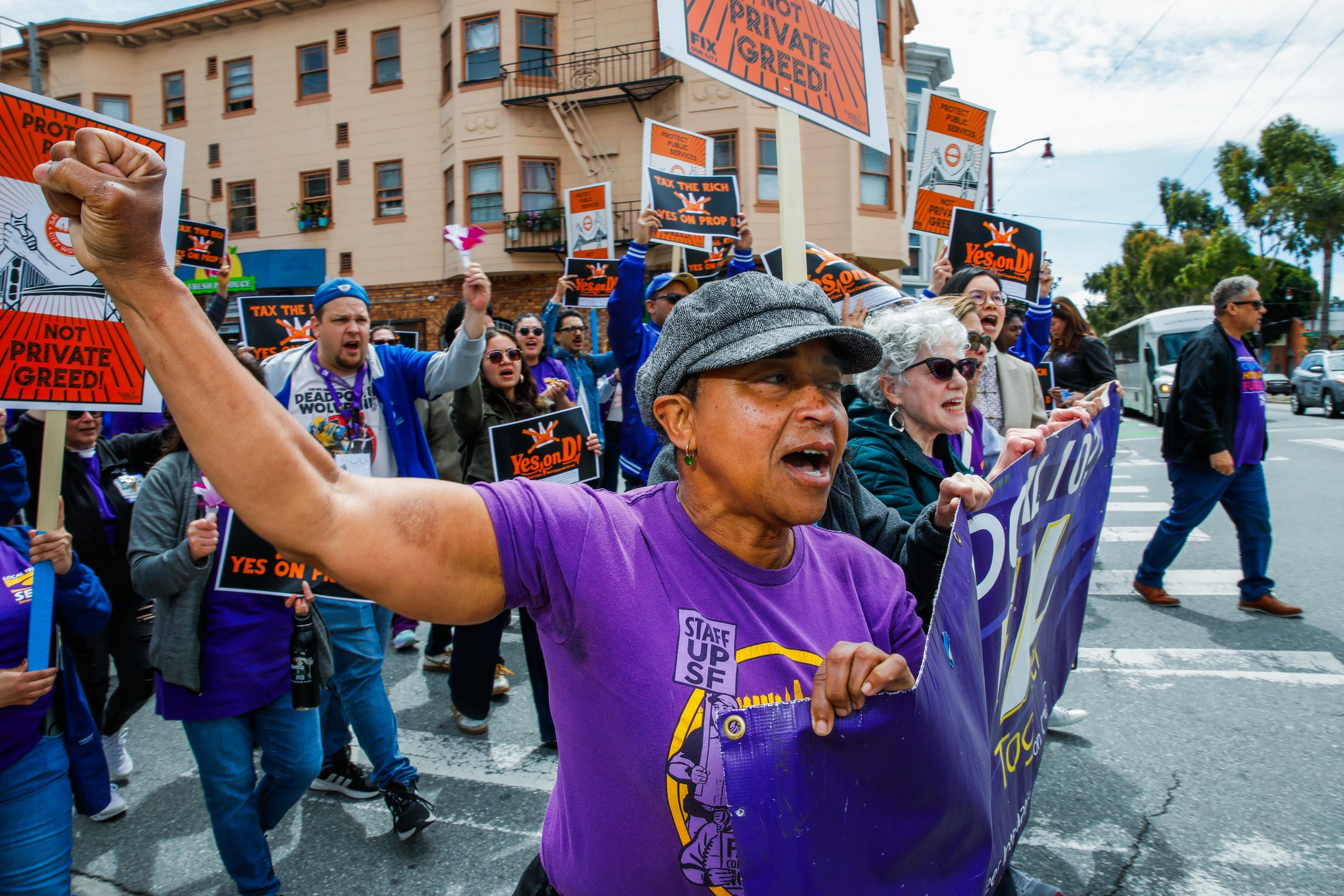 A diverse group of protesters march on a city street, holding signs advocating "Yes on Prop 15" and chanting slogans against private greed.