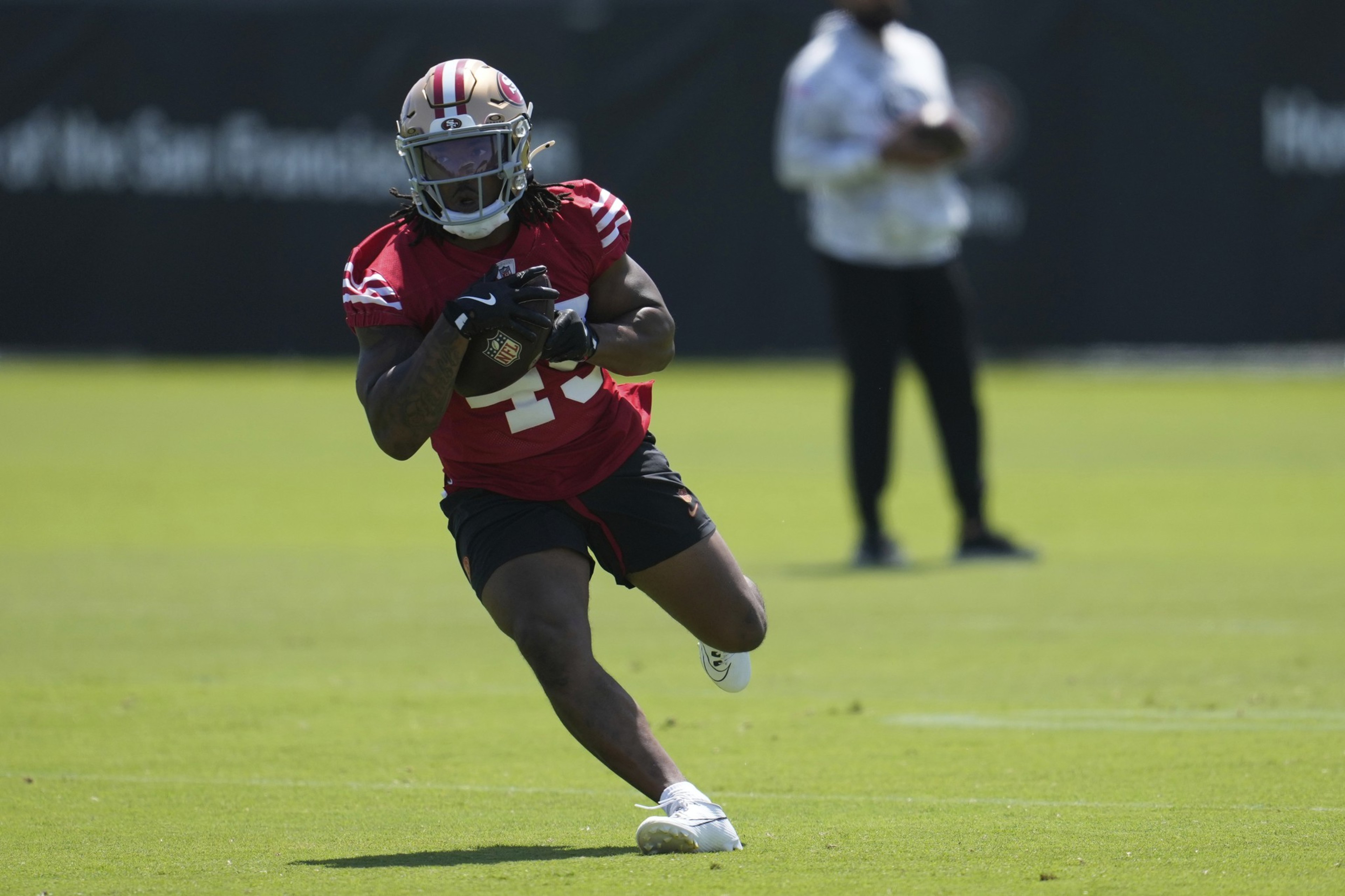 A football player in a red jersey, numbered 23, runs on a grassy field, holding a ball. He's wearing a helmet and gloves, with a person blurred in the background.