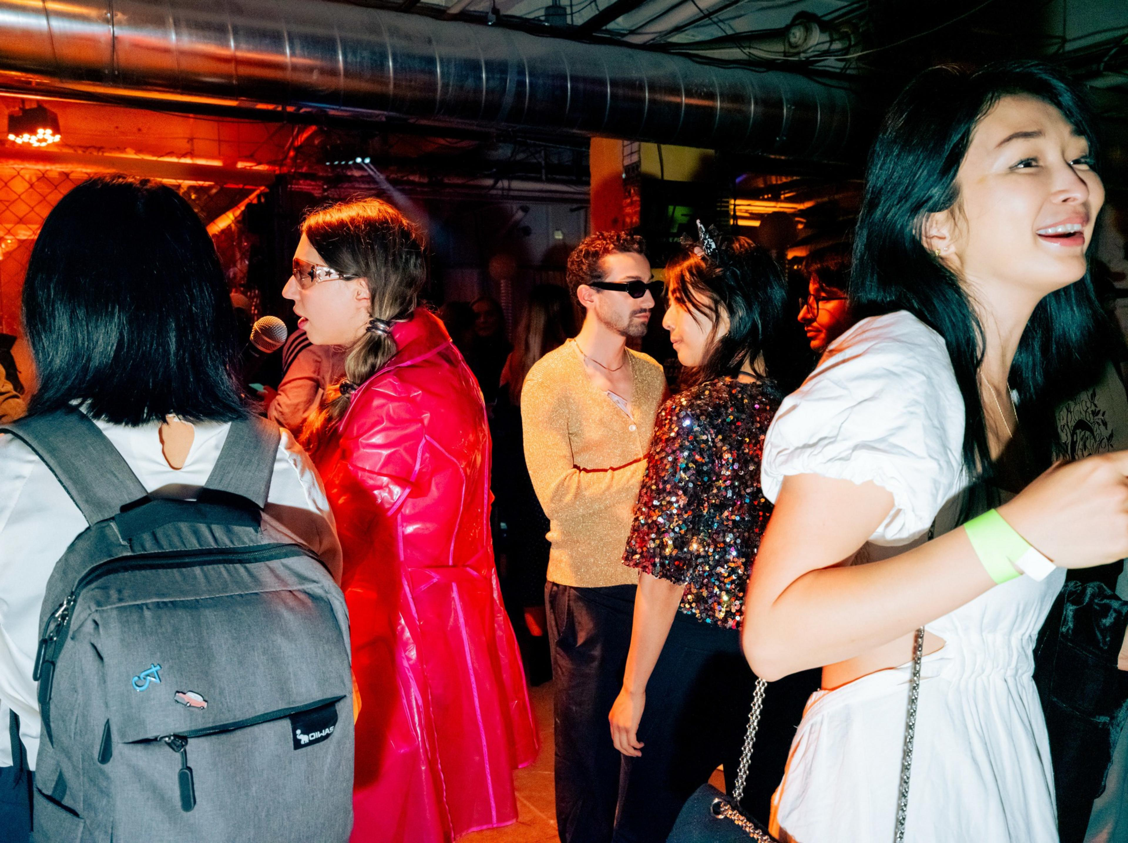 A group of people wearing colorful, stylish outfits socialize in a dimly lit room with exposed pipes and an industrial vibe.