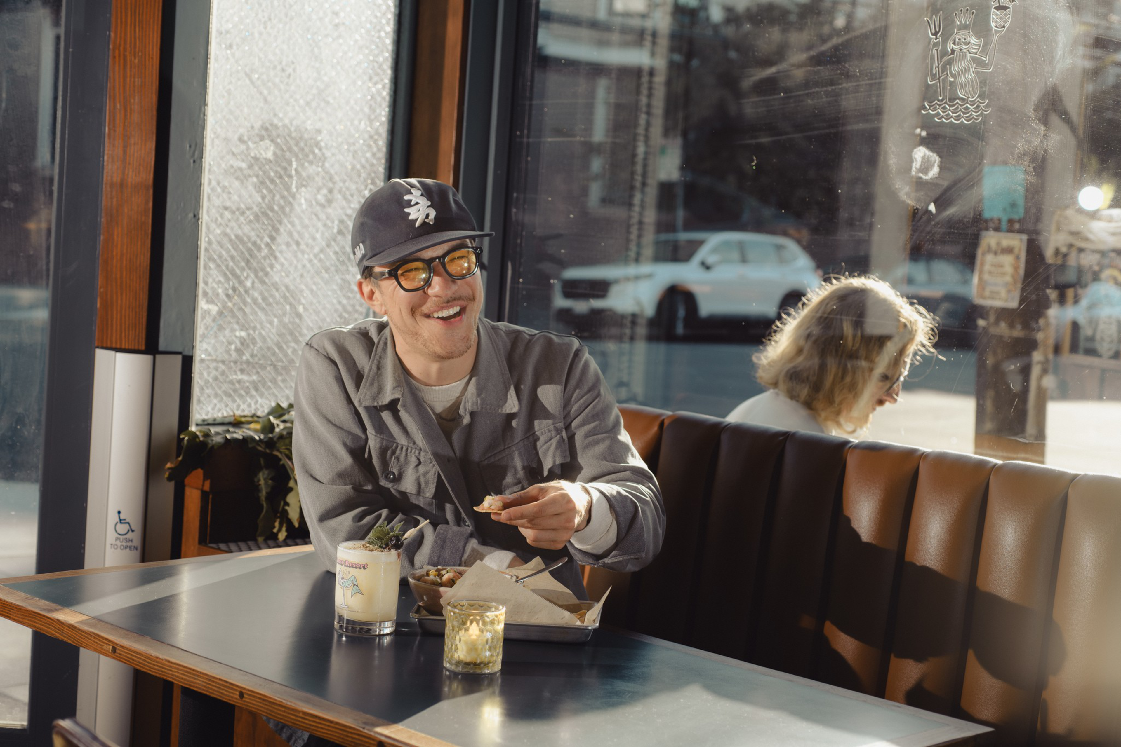A man wearing glasses and a cap smiles while eating at a table with a drink and candle, sunlight streaming through large windows behind him.