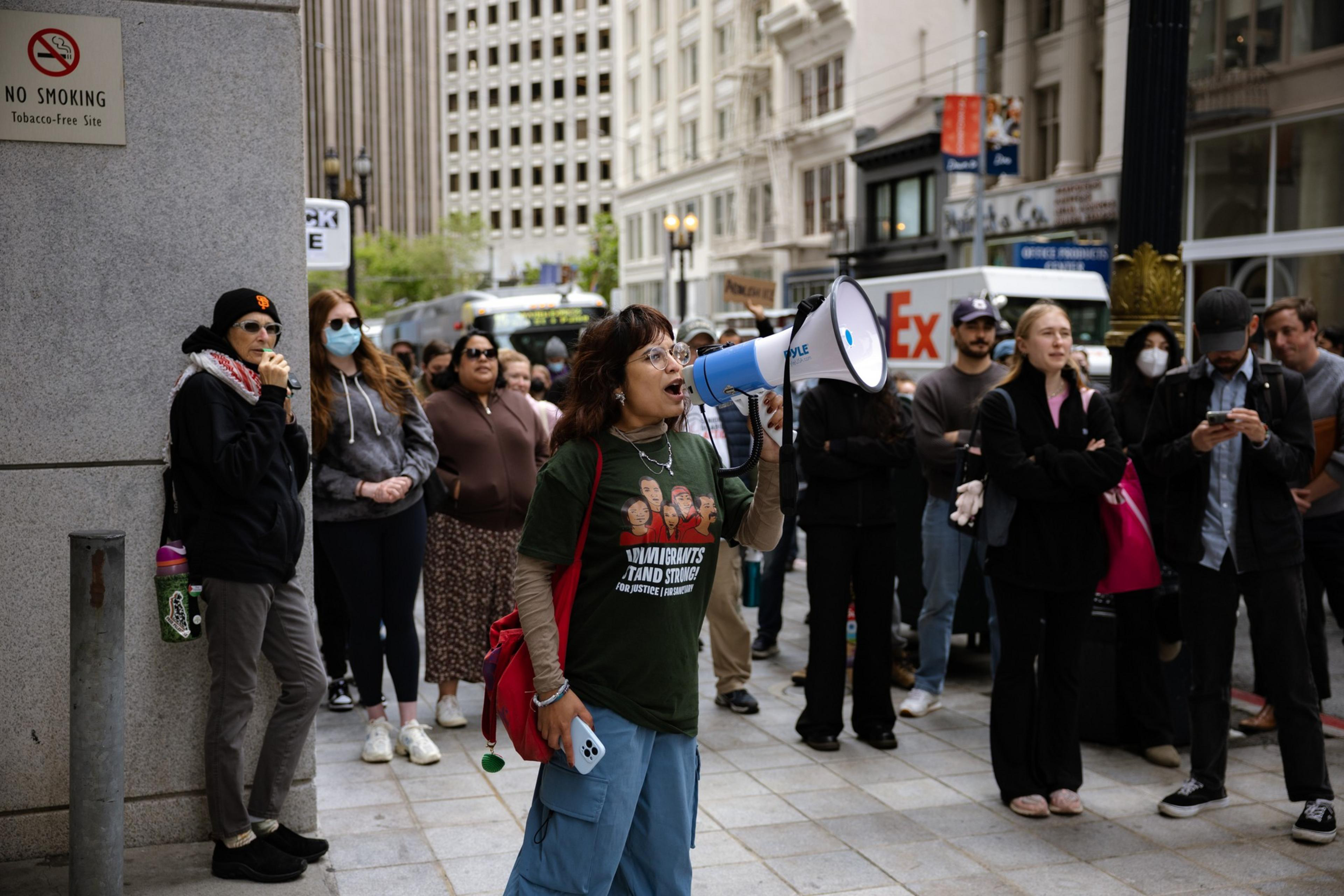 A person uses a megaphone, leading a protest or rally. A diverse group stands in the background, some with masks, observing in a city street setting.