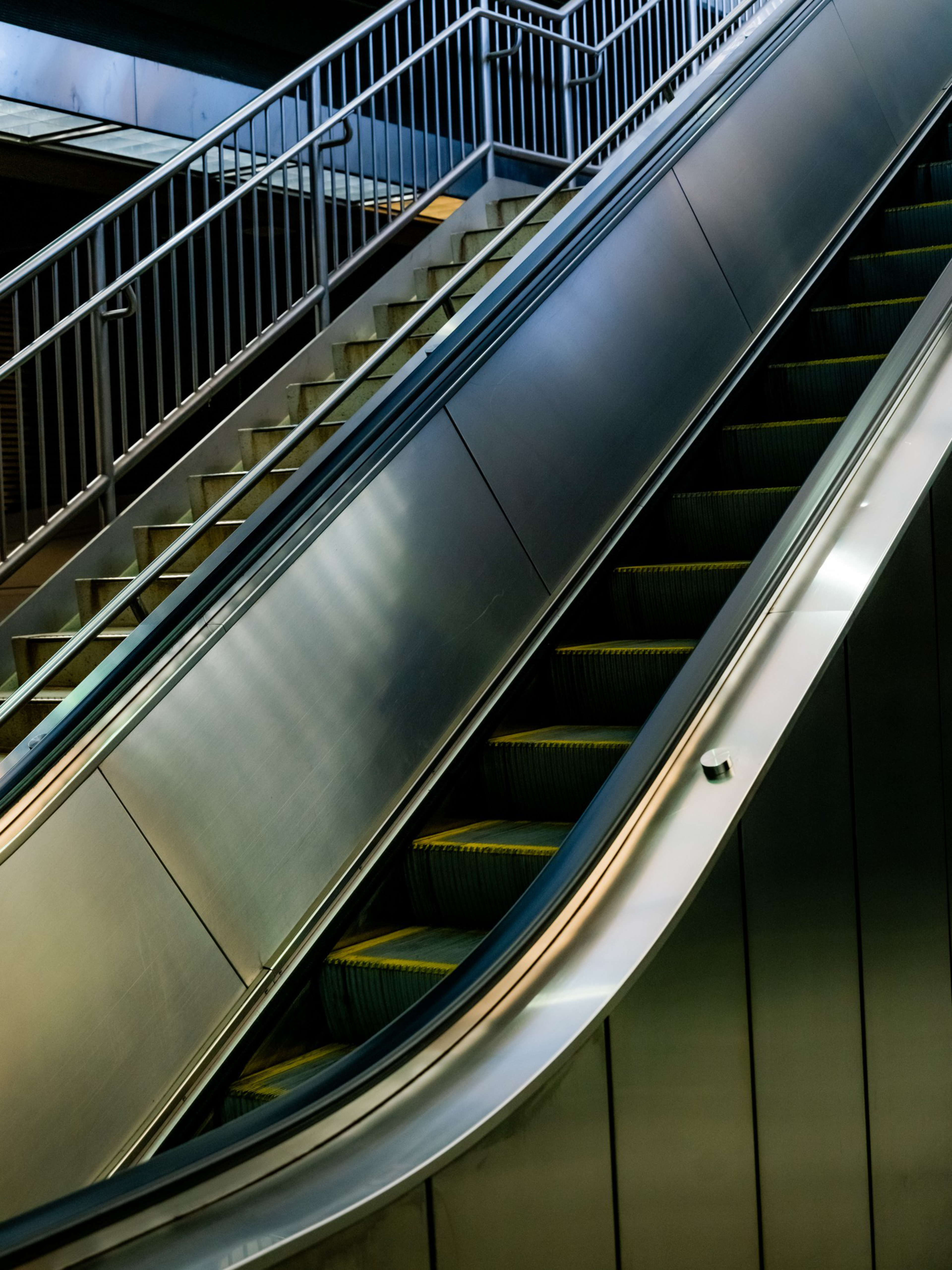 A descending escalator runs parallel to a stationary stairway with metal railings, all set in a modern indoor environment.