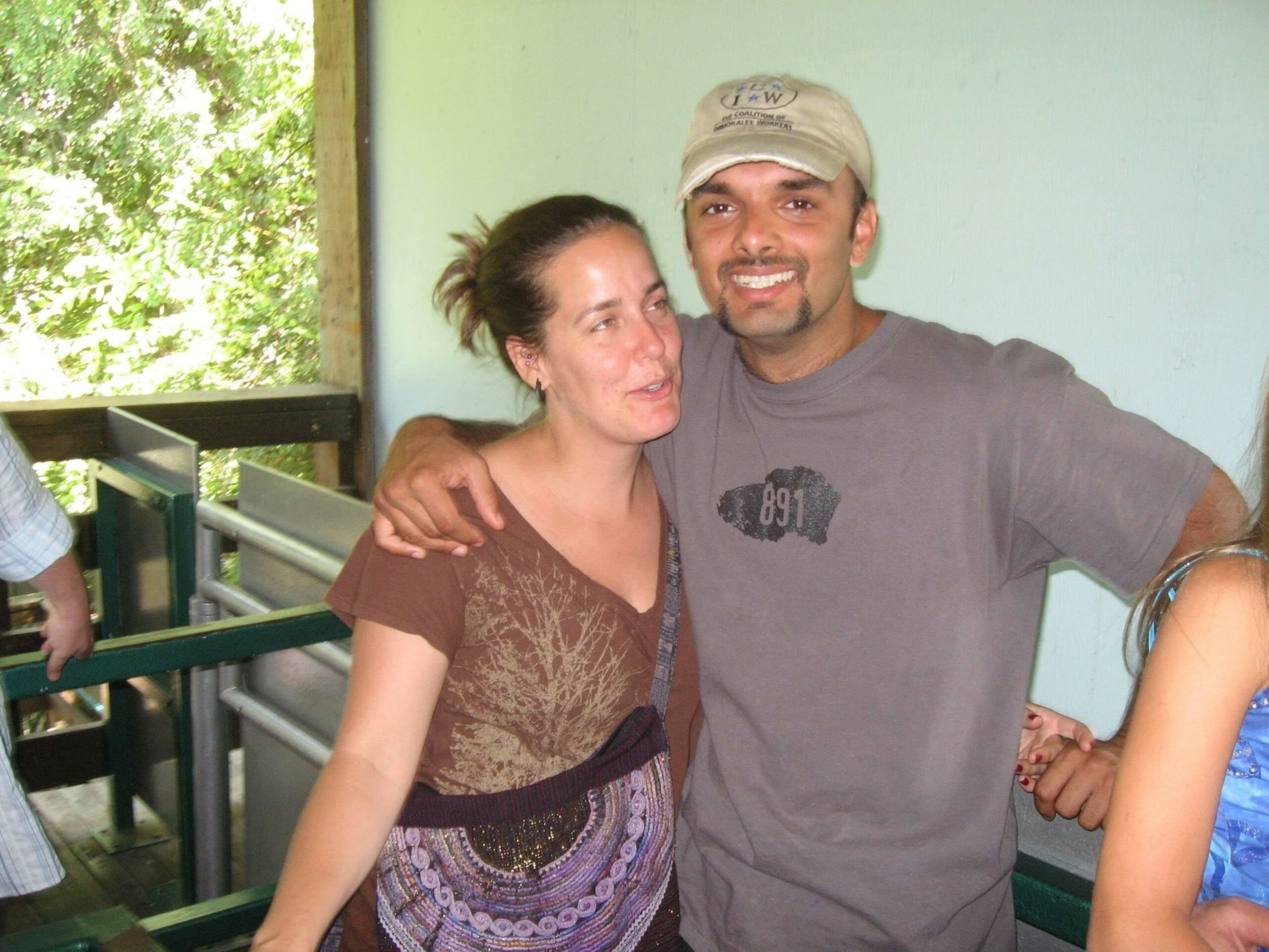 A man in a beige cap and gray shirt smiles with his arm around a woman in a brown shirt with a tree design, standing indoors near a balcony.