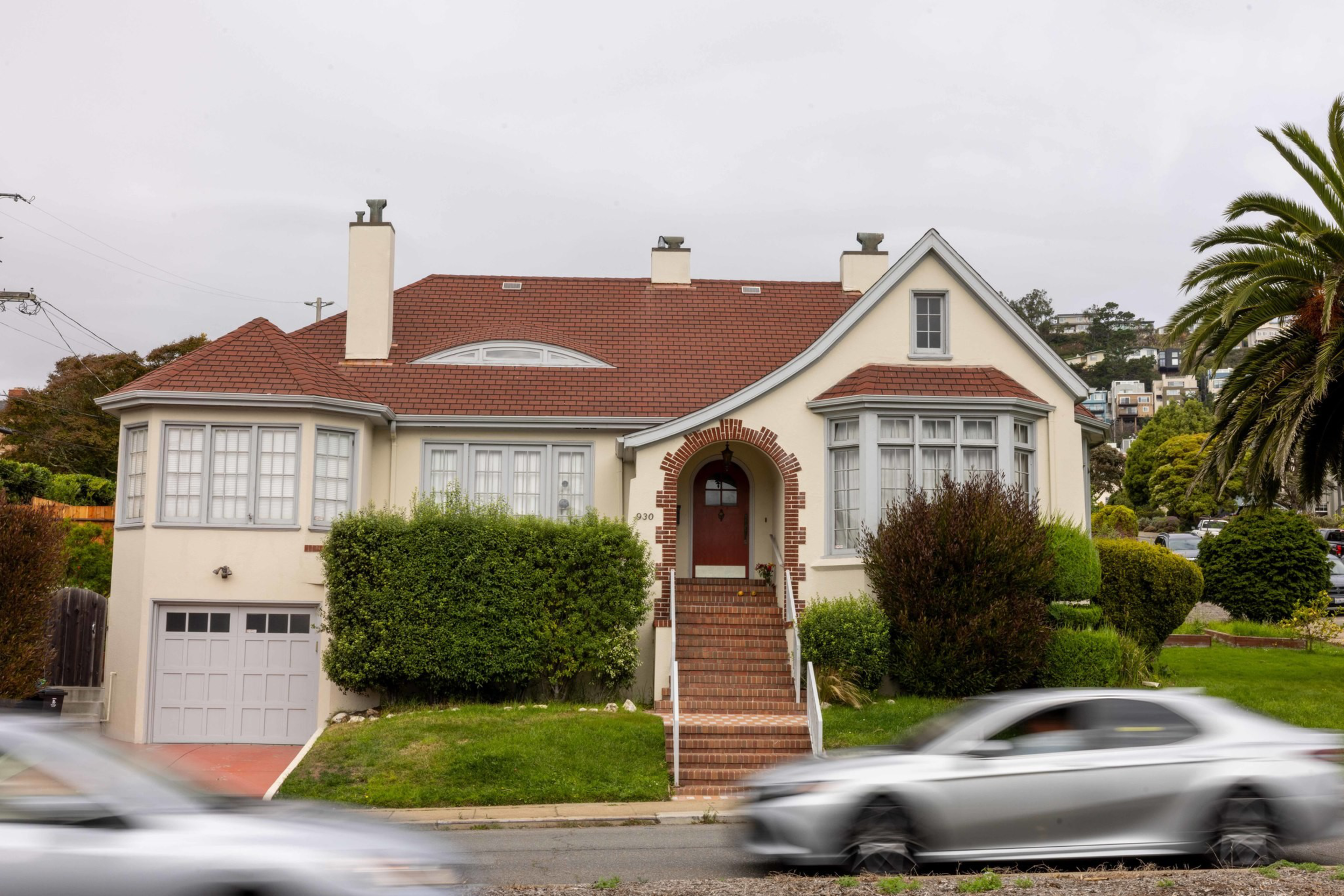 A beige house with a red-tiled roof features a central arched entrance reached by brick stairs, surrounded by bushes, with cars driving past on the street.