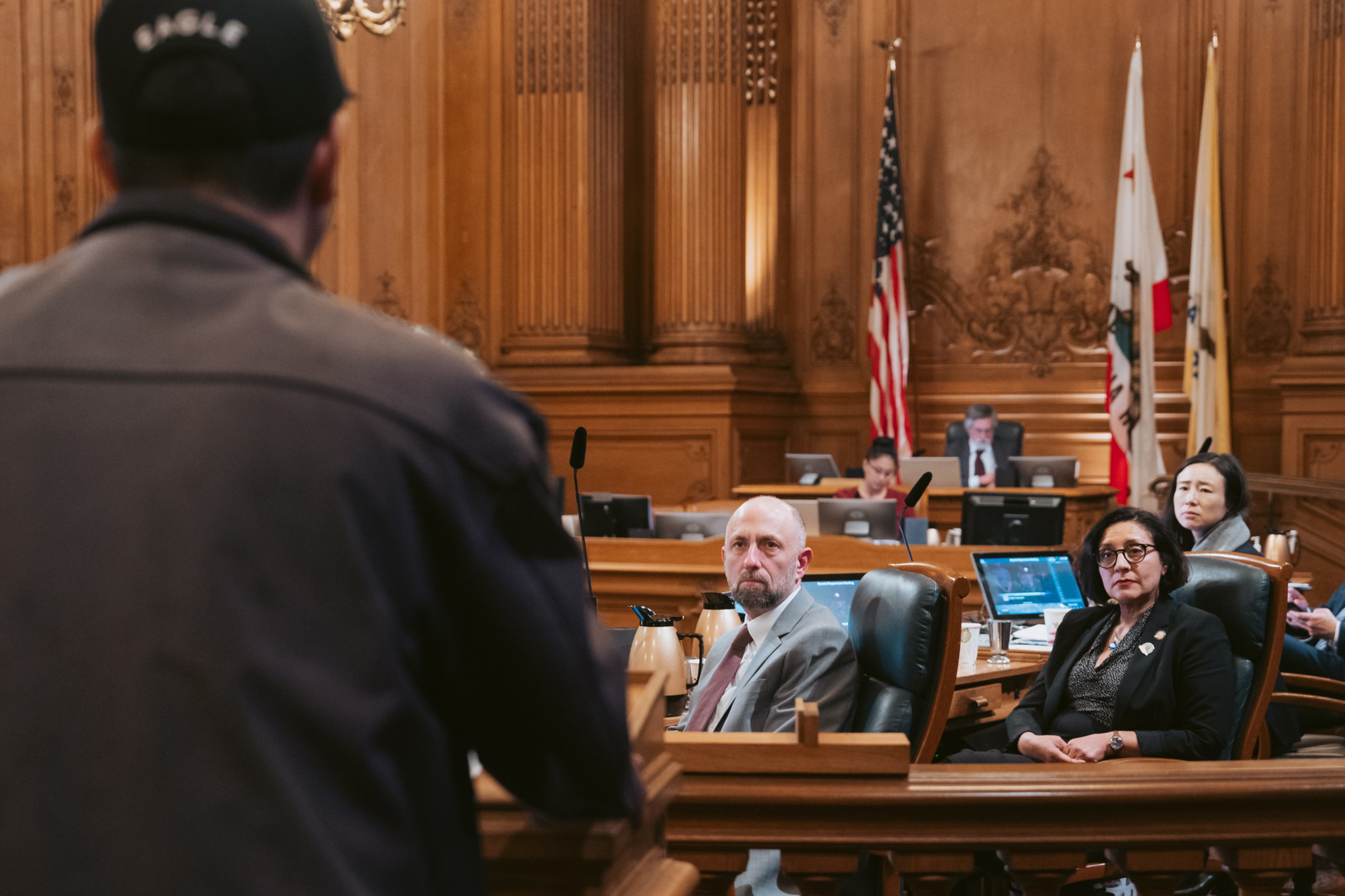 A man in a hat and jacket speaks in a wood-paneled council chamber to seated officials, including two women and one man, with flags in the background.