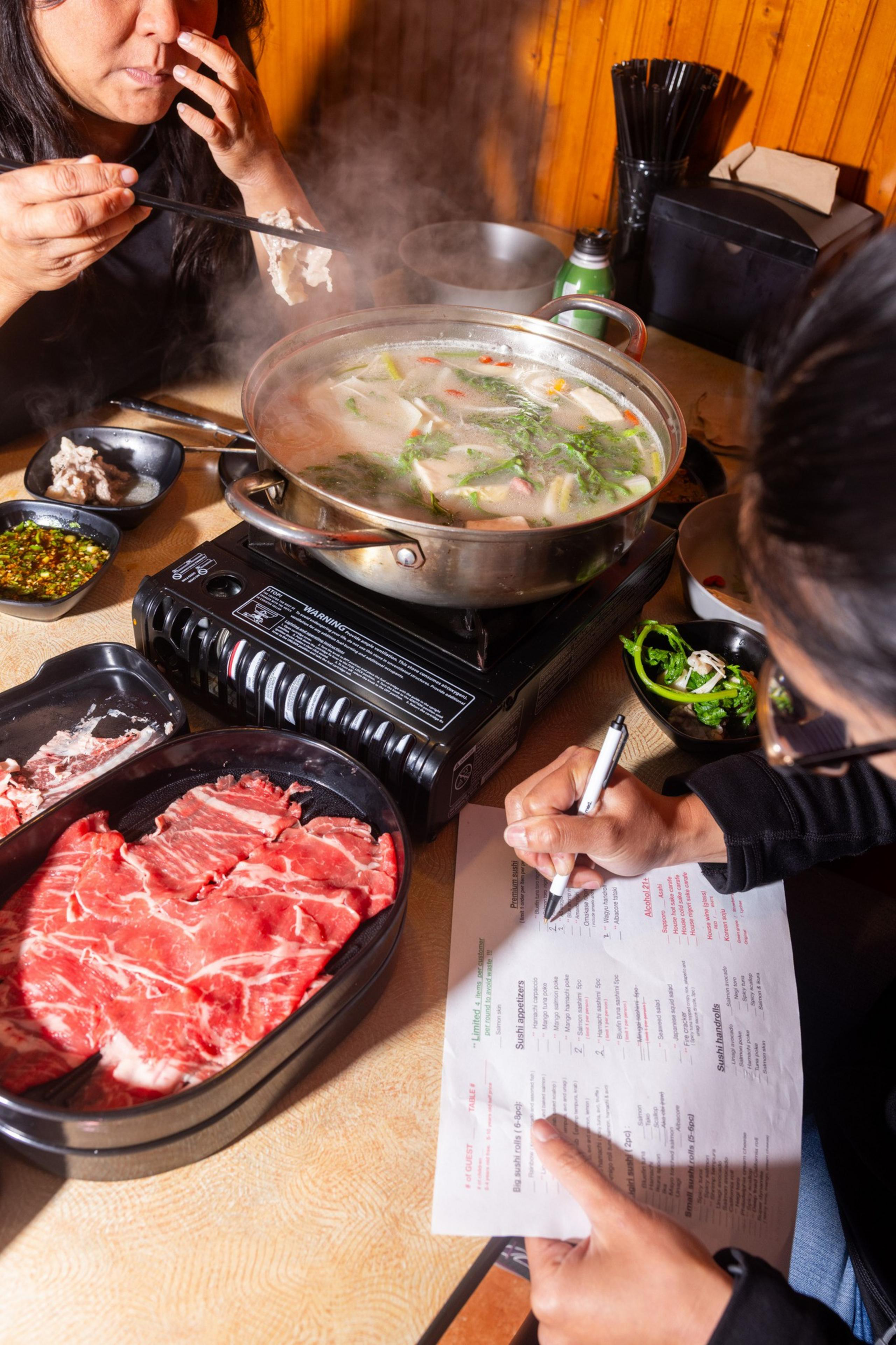 Two people enjoy a hot pot meal. One person uses chopsticks, while the other writes on a menu. A pot of steaming broth and raw meat are on the table.
