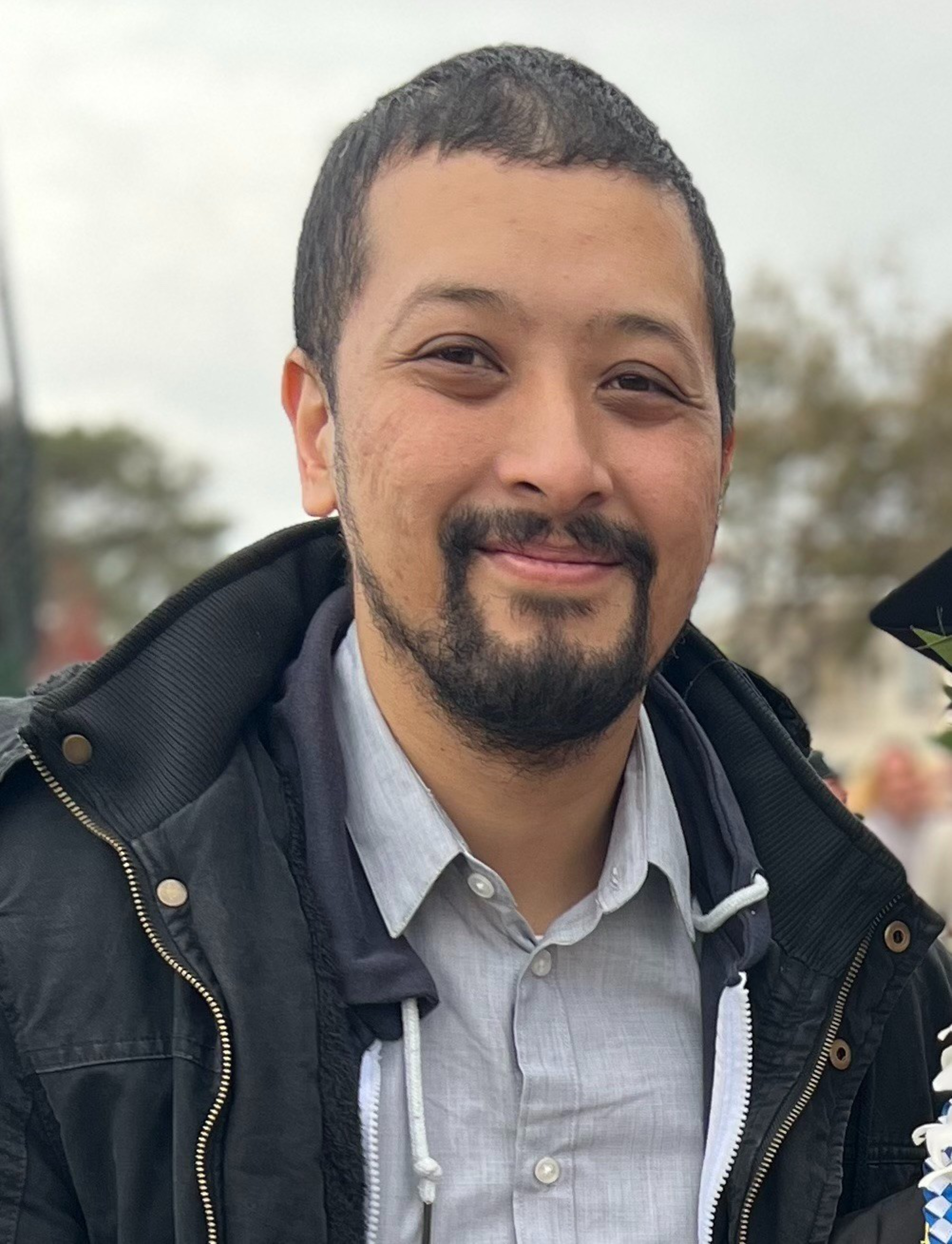 A man with short dark hair and a beard smiles gently, wearing a grey button-up shirt under a black jacket with a hoodie.