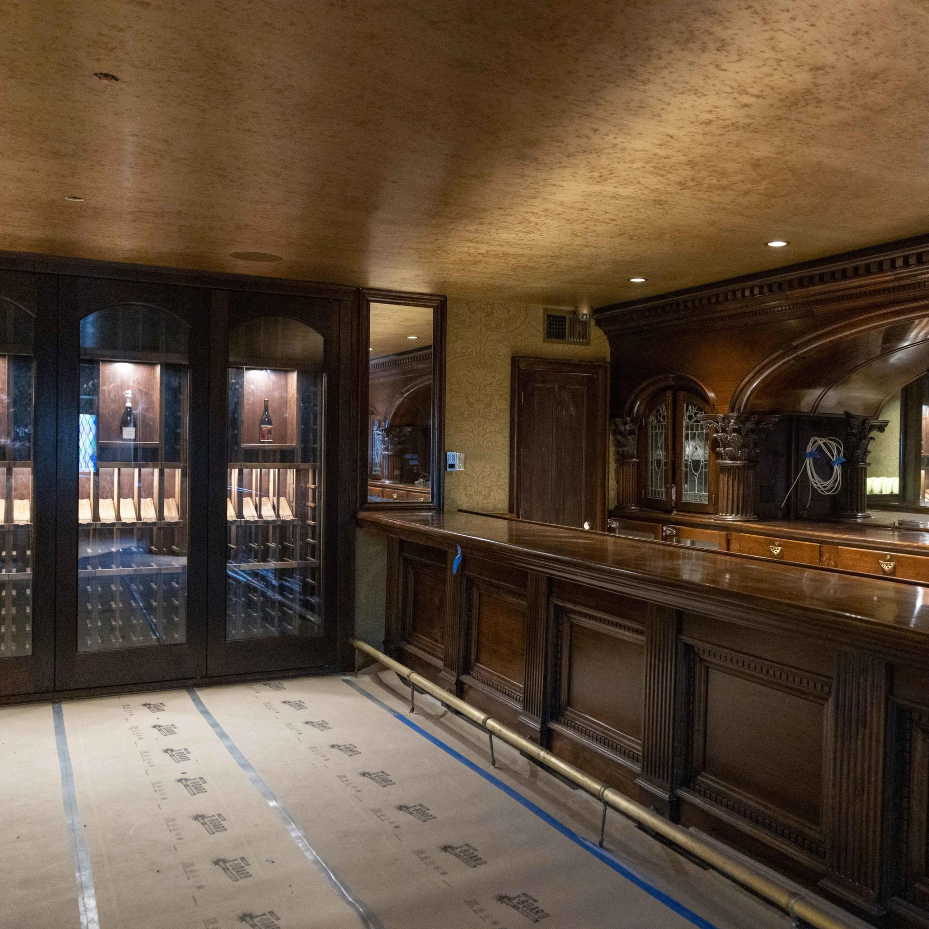 The image shows a room with rich, dark wood paneling, a long wooden bar, and a wine display cabinet with glass doors. The floor is covered with protective paper.
