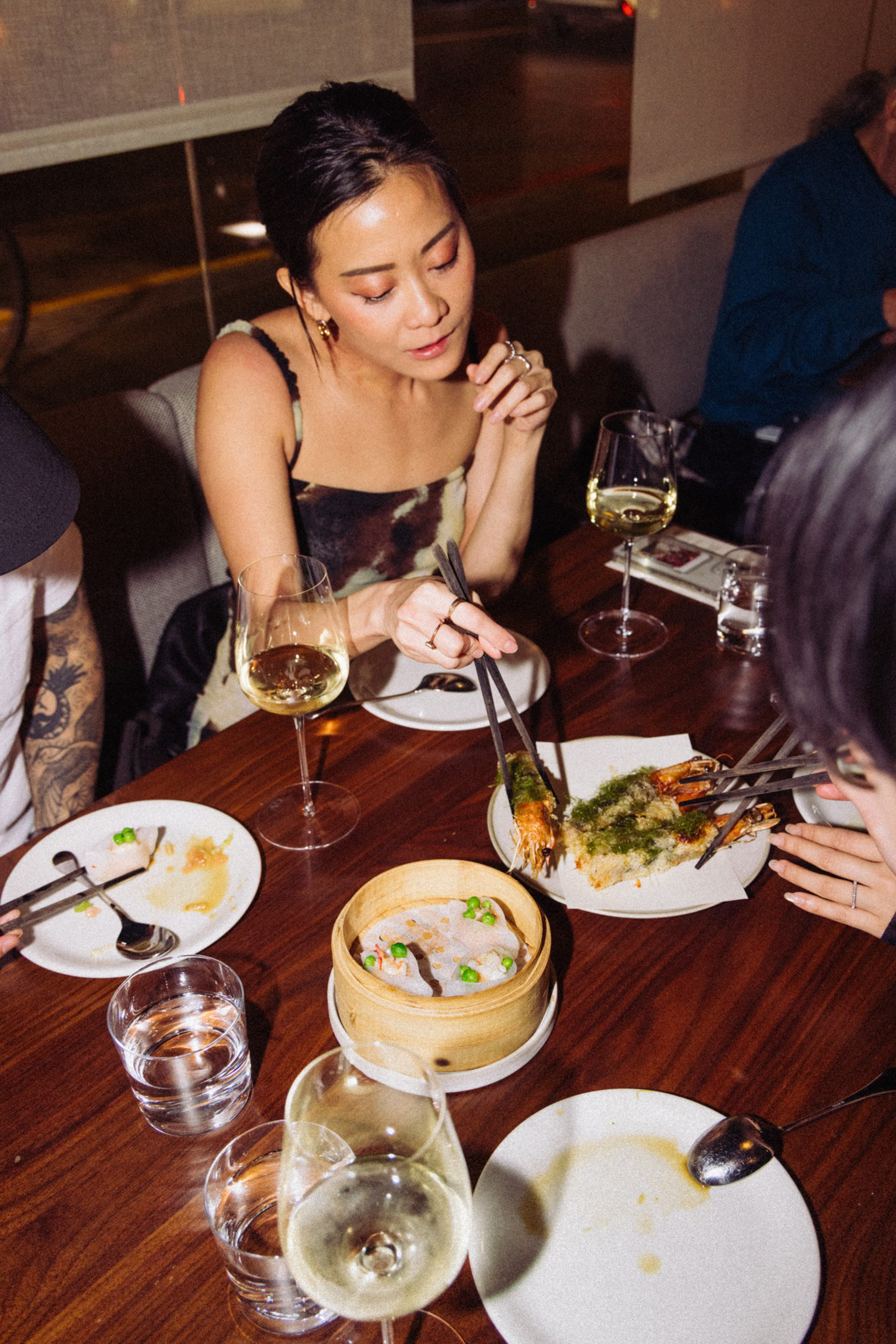 A woman uses chopsticks to pick up food from a plate of shrimp while sitting at a table with wine glasses, water, and a bamboo steamer basket.