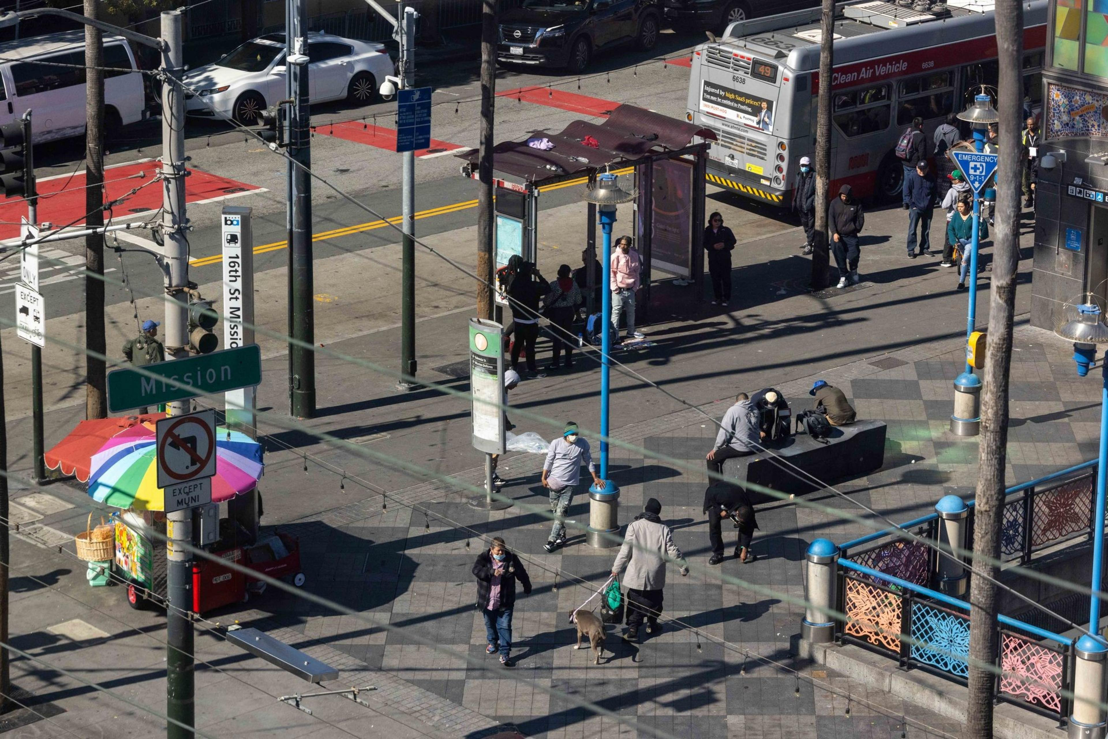 People are gathered at a city intersection, near a colorful street vendor cart and a bus stop. A few are seated, some are walking, and a bus is parked nearby.