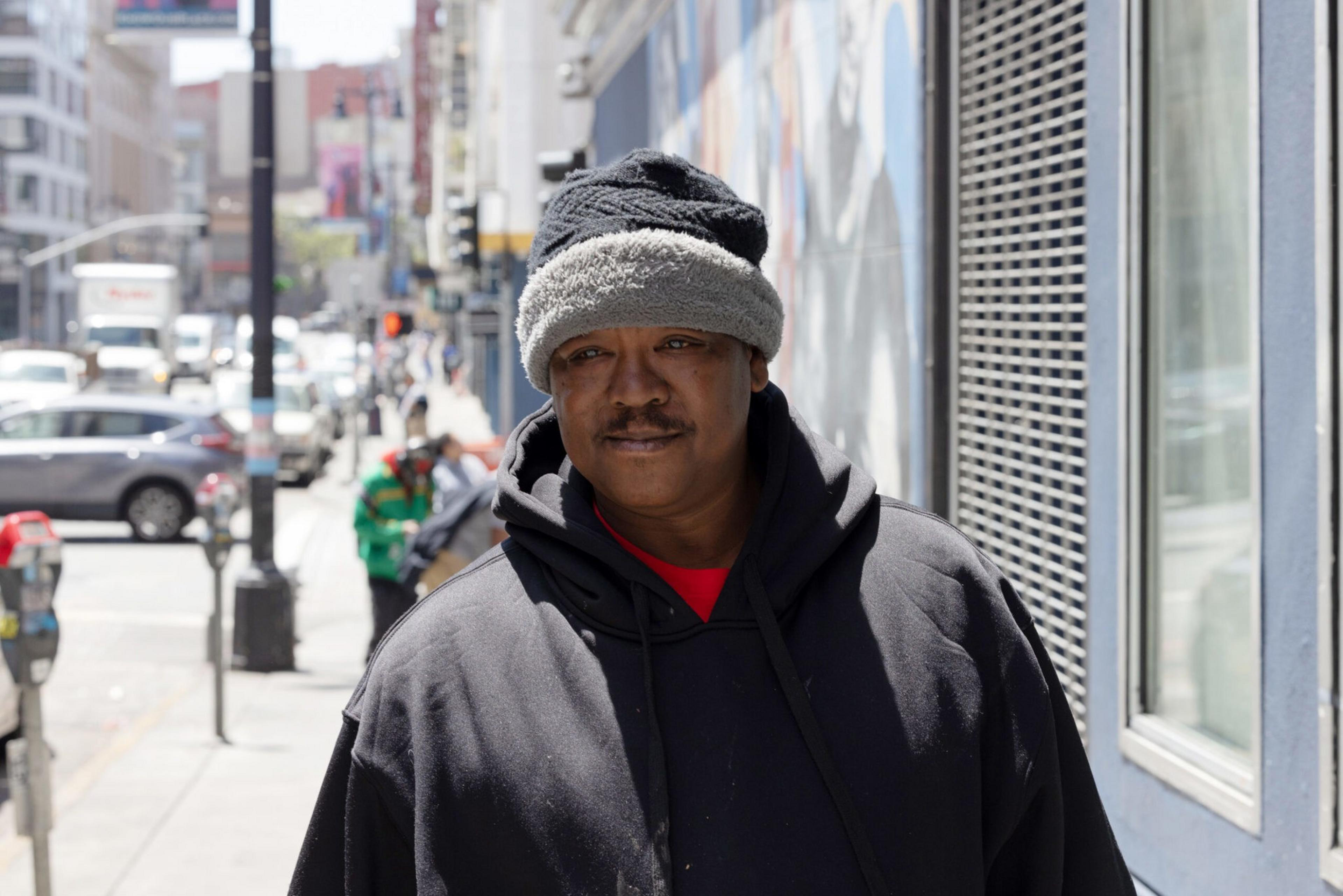 A person wearing a dark hoodie and a gray, fuzzy hat stands on a busy city sidewalk, with several cars and pedestrians visible in the background.