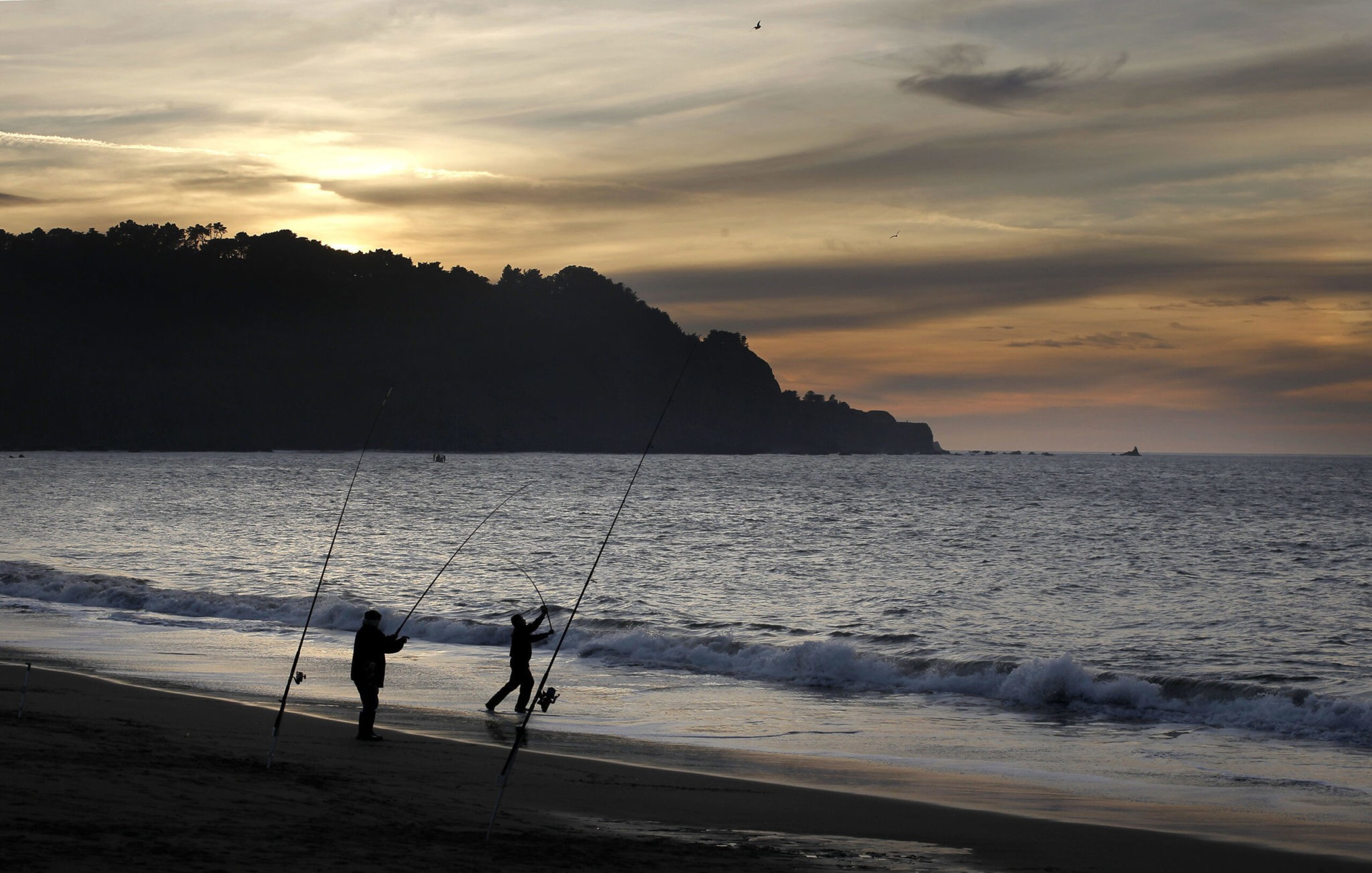 Two people are fishing on a beach at sunset, with fishing rods planted in the sand and waves gently rolling onto the shore.