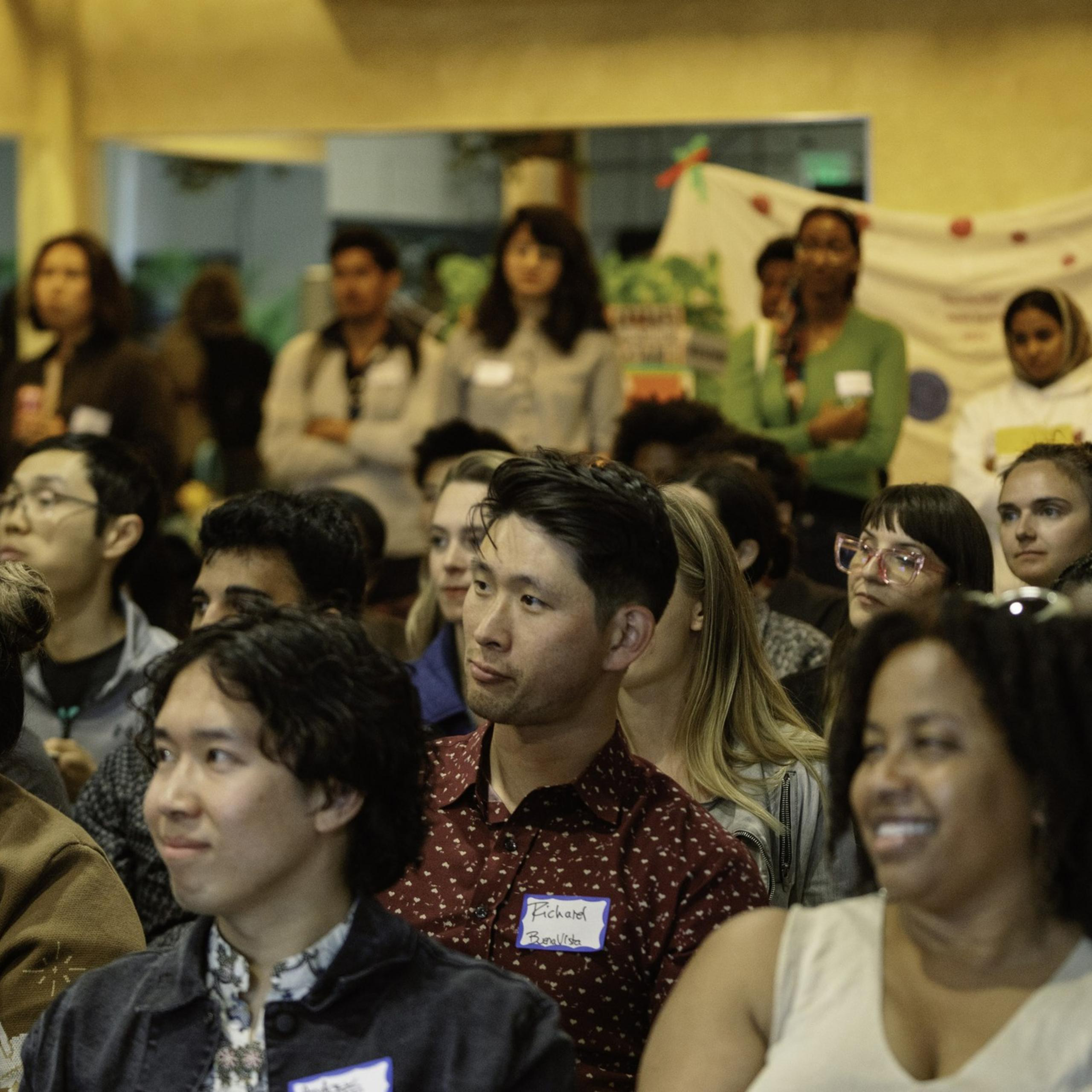 A diverse group of people attentively watch something out of the frame in an indoor setting, with a mix of sitting and standing individuals. Some wear name tags.