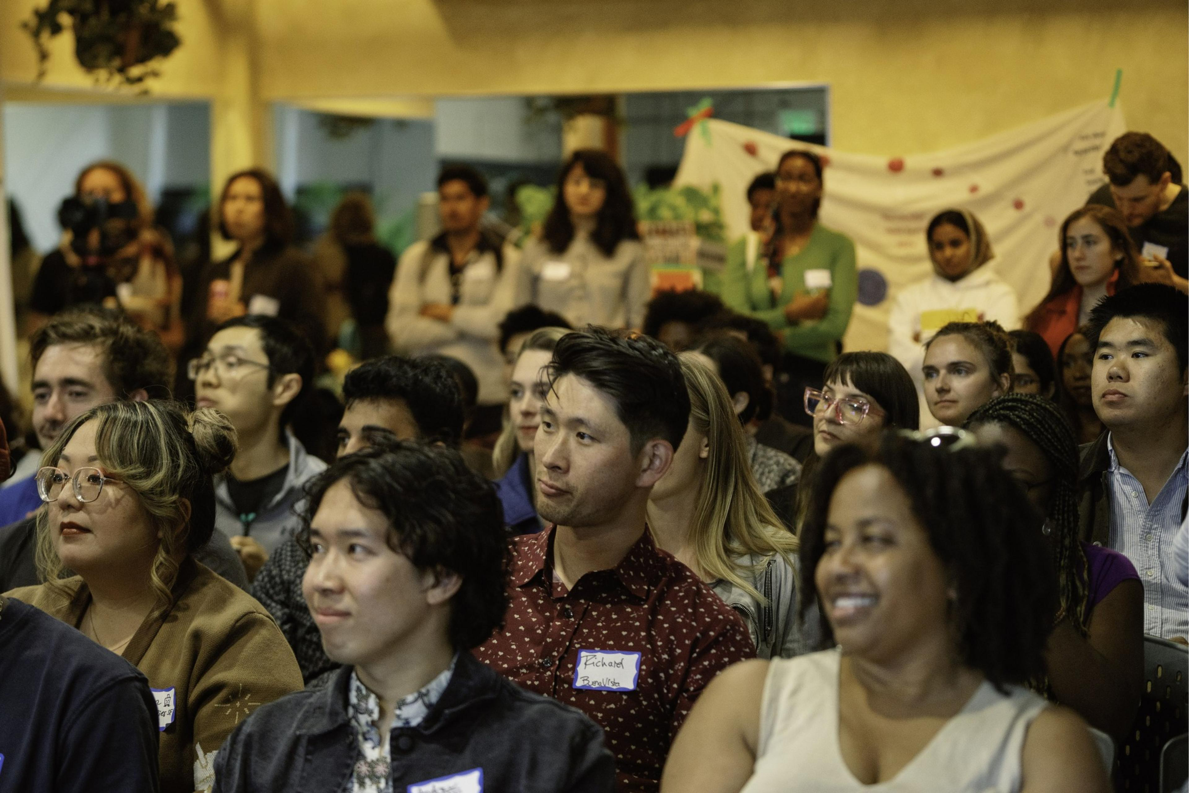 A diverse group of people attentively watch something out of the frame in an indoor setting, with a mix of sitting and standing individuals. Some wear name tags.