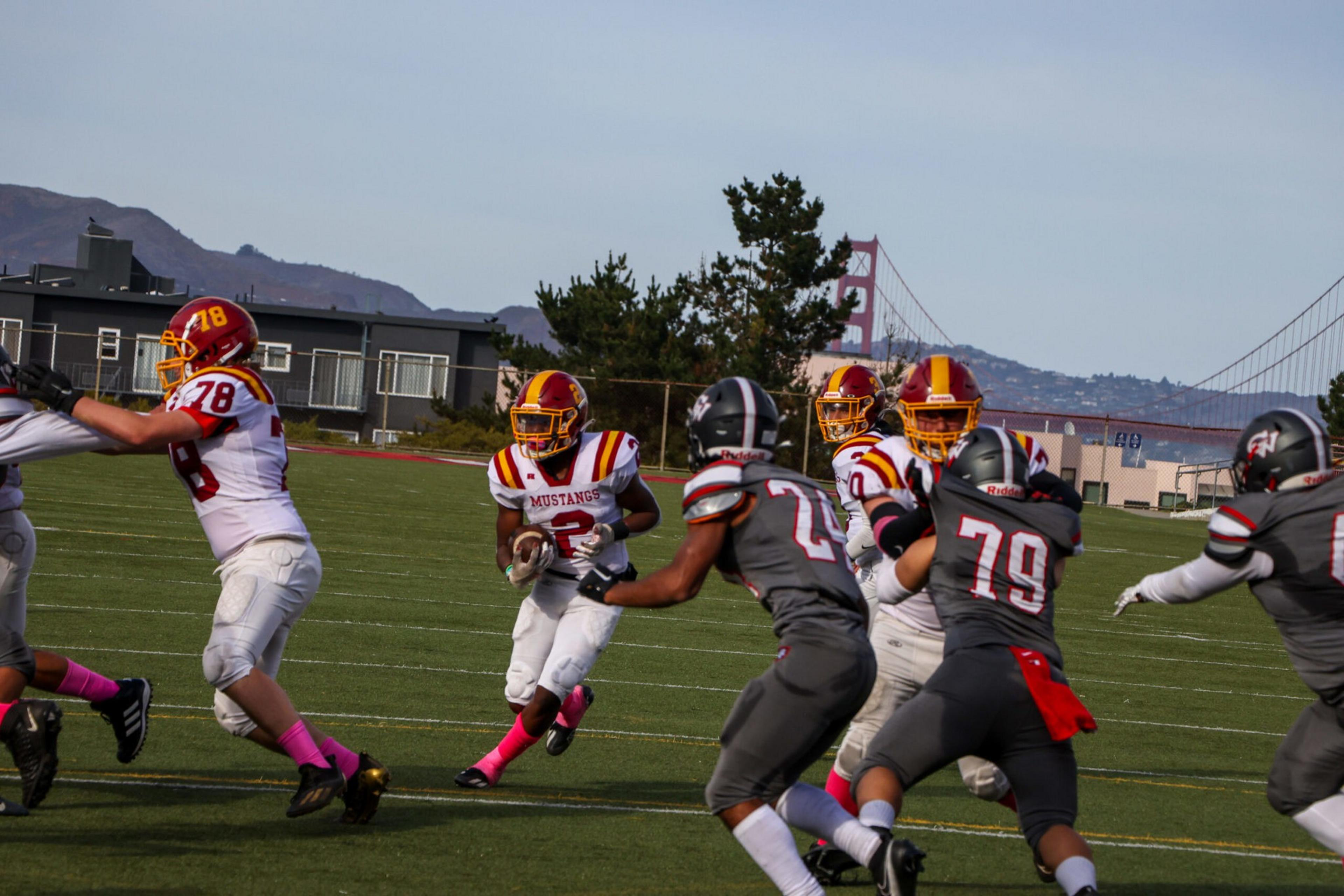 A football player in a white and red Mustangs uniform runs with the ball, while players in gray uniforms attempt to block or tackle him on a green field.