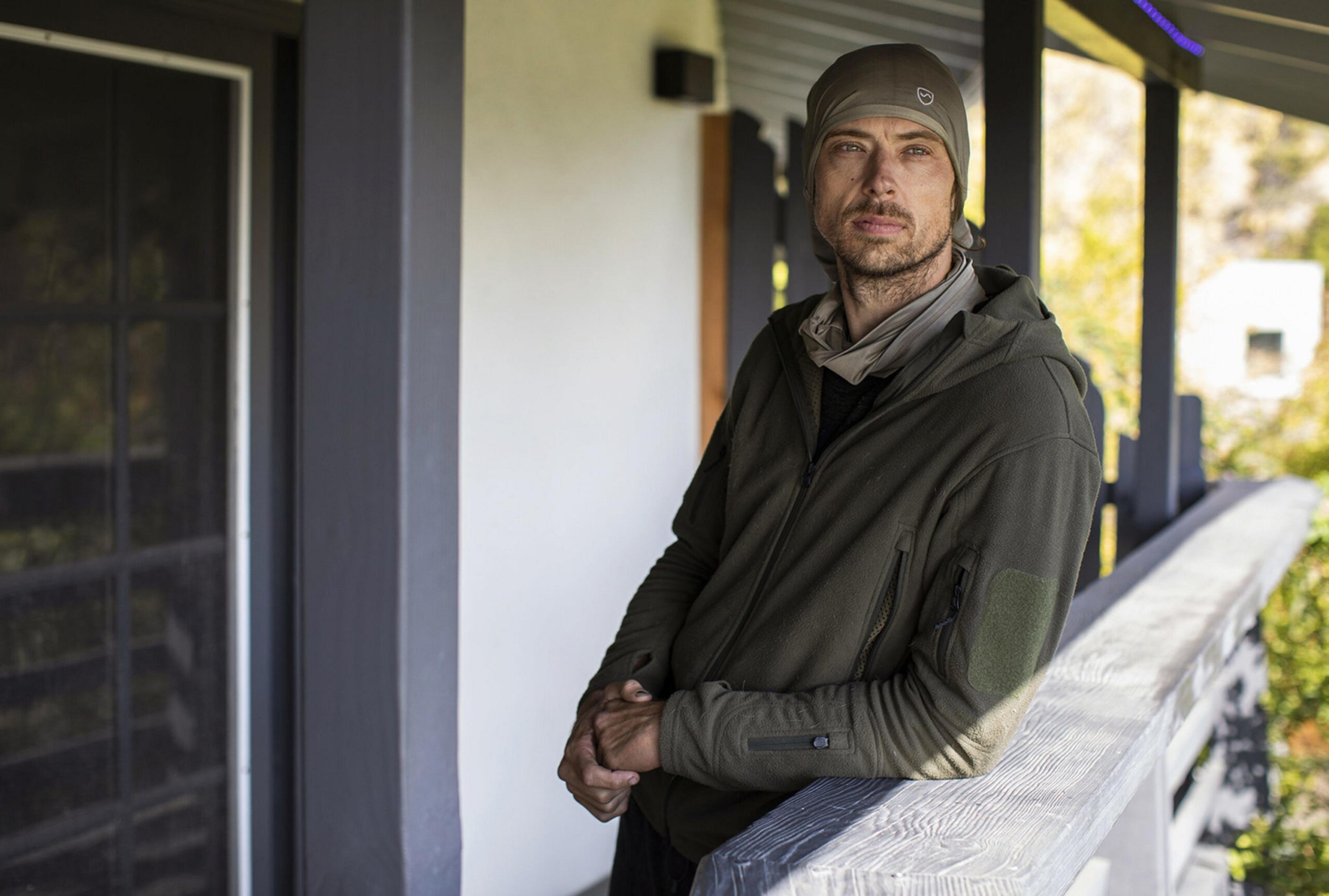 A man wearing a green hoodie and a matching beanie leans on a railing outside a building, looking directly at the camera with a calm expression.