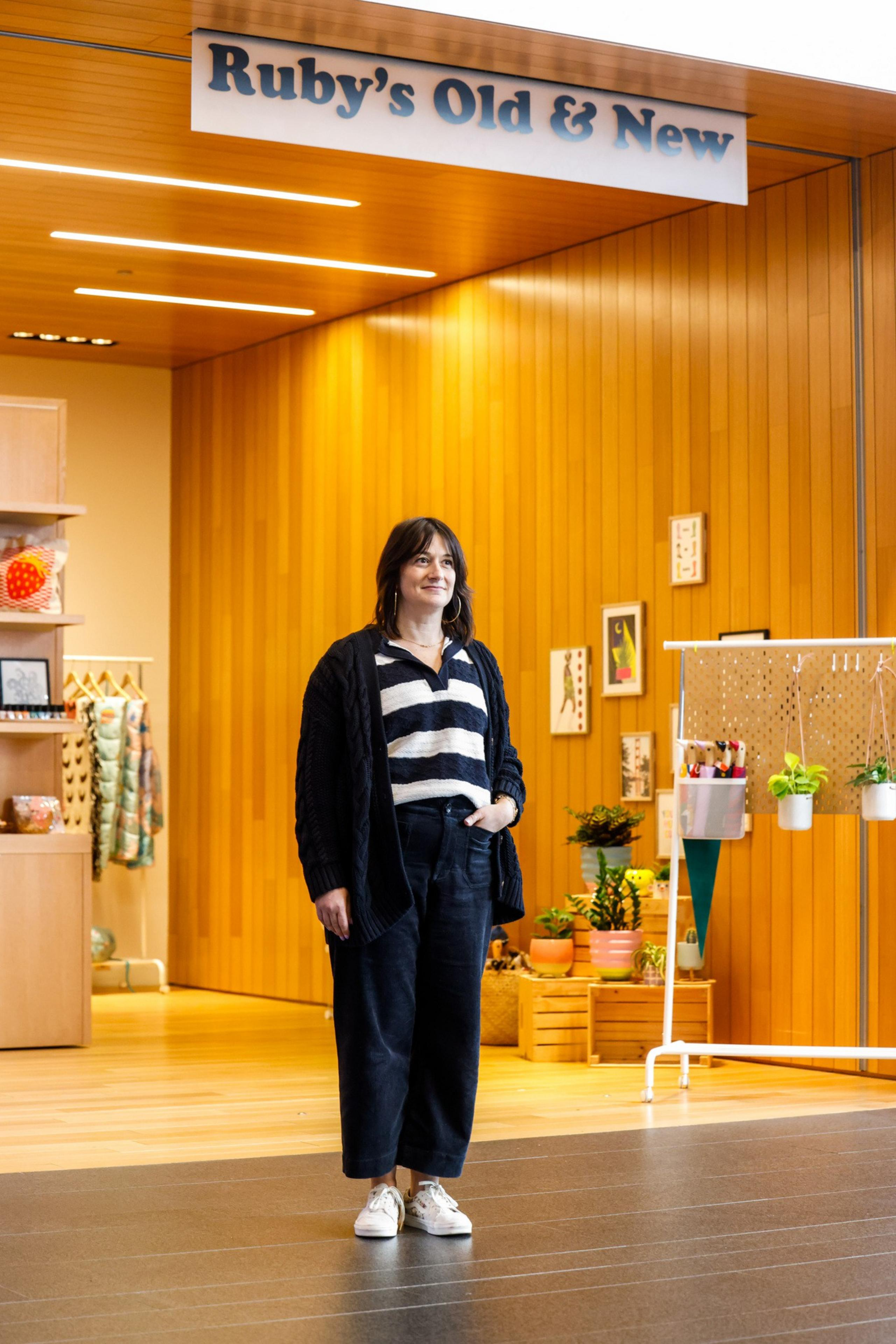 A person stands in front of a store called &quot;Ruby's Old &amp; New,&quot; featuring wood paneling, plants, clothes, and framed art. The person is wearing casual attire.