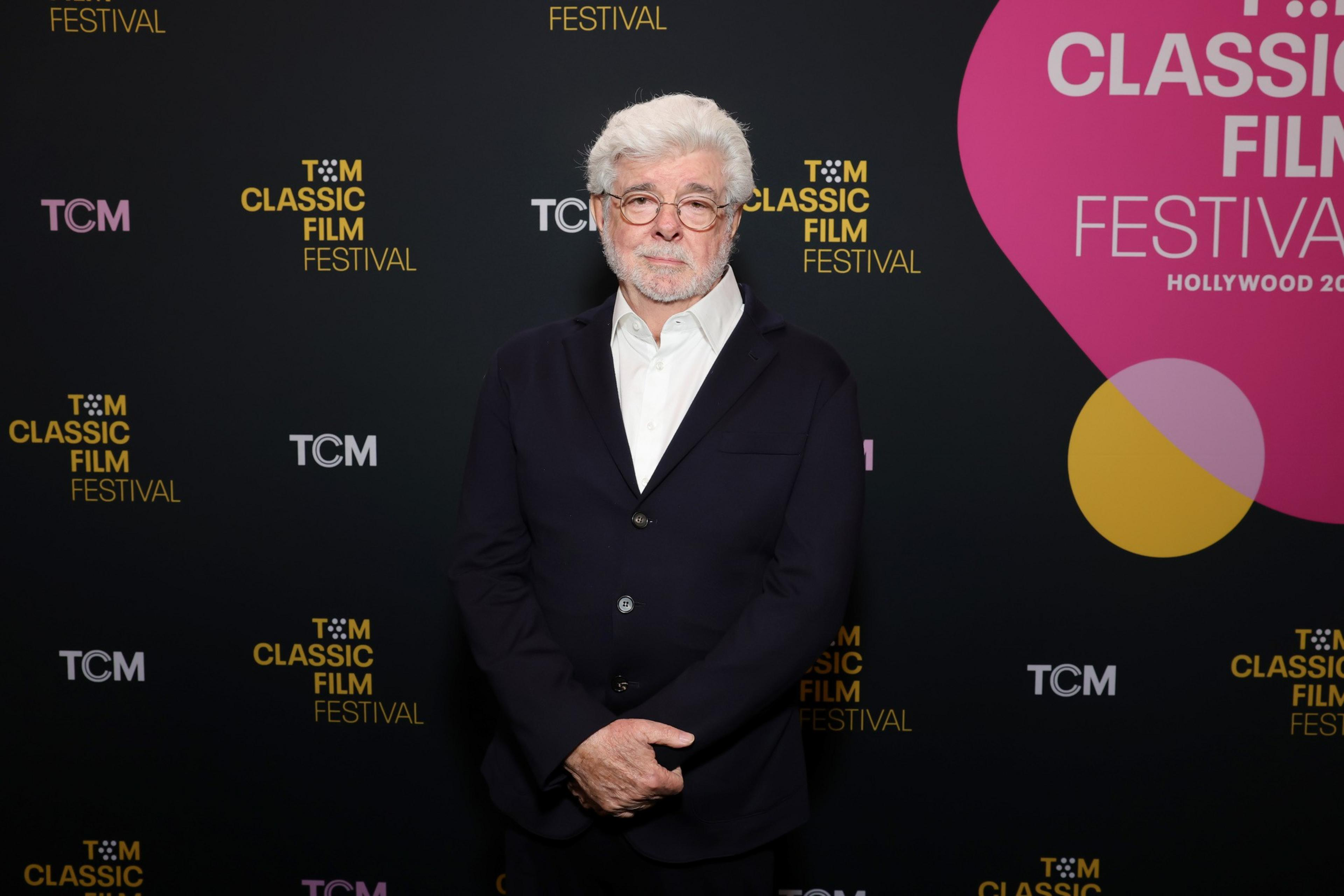 A man with white hair and glasses stands against a backdrop with &quot;TCM Classic Film Festival&quot; logos, wearing a dark suit and white shirt.