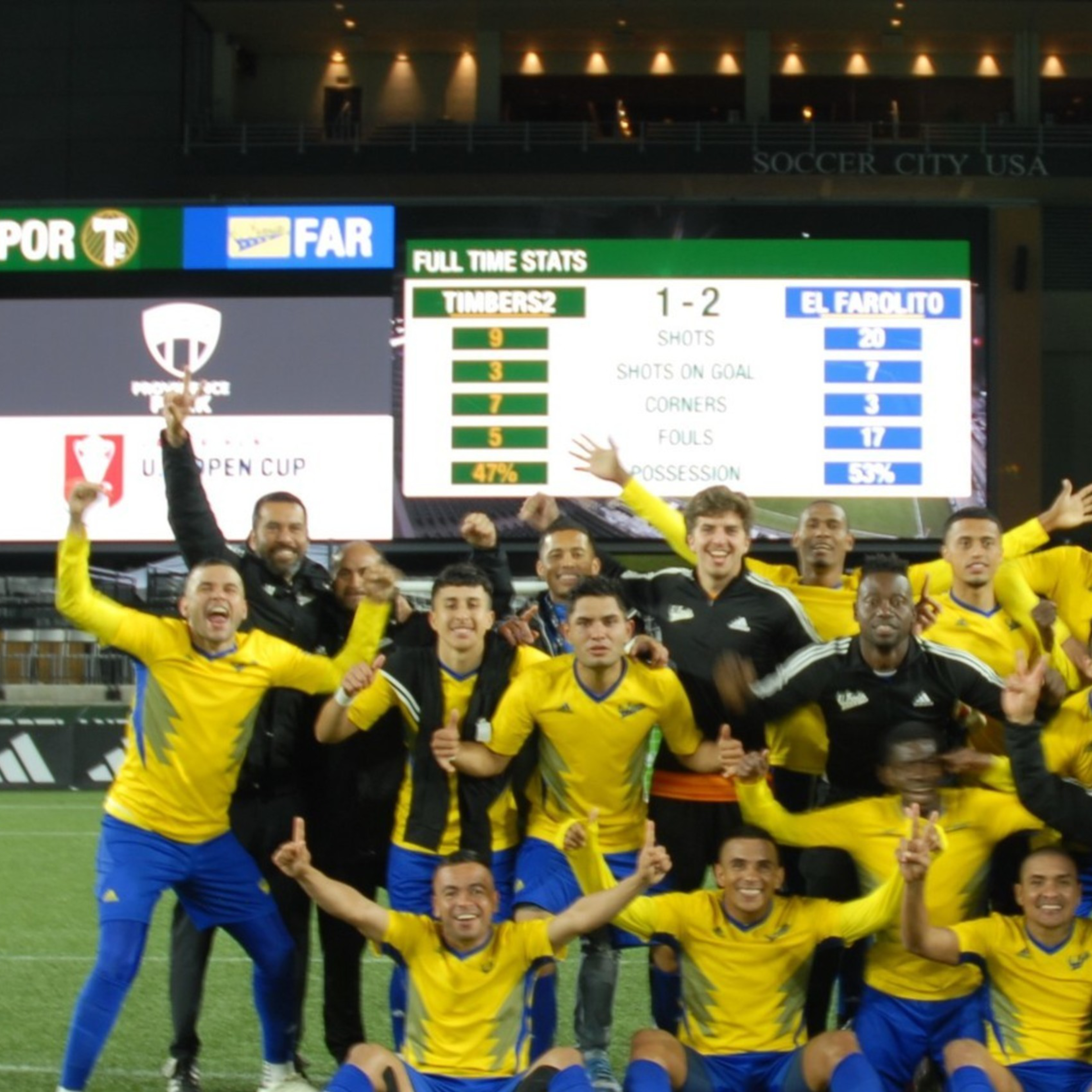 A jubilant soccer team celebrates on a pitch at night with a scoreboard showing a 1-2 victory in the background.