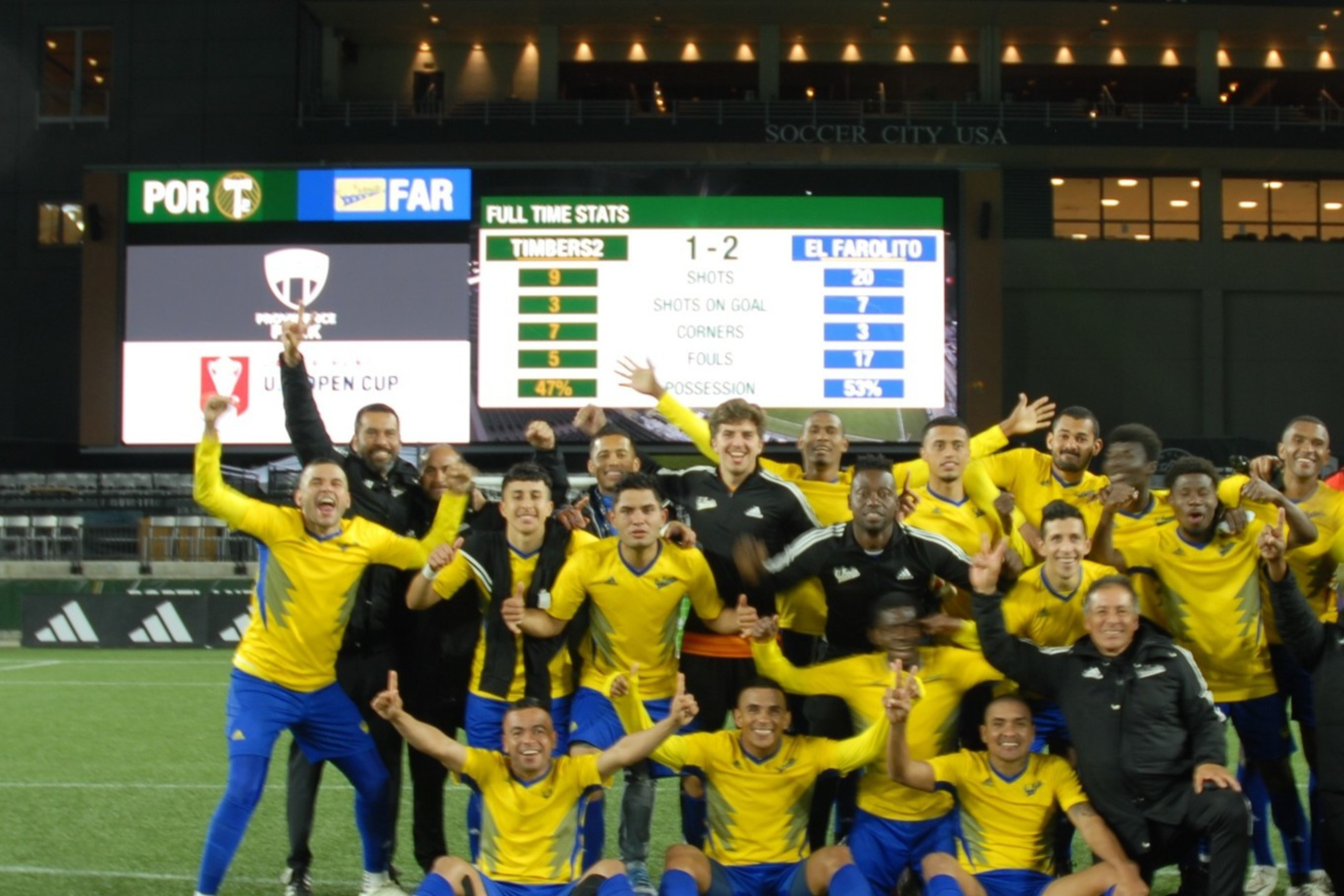 A jubilant soccer team celebrates on a pitch at night with a scoreboard showing a 1-2 victory in the background.