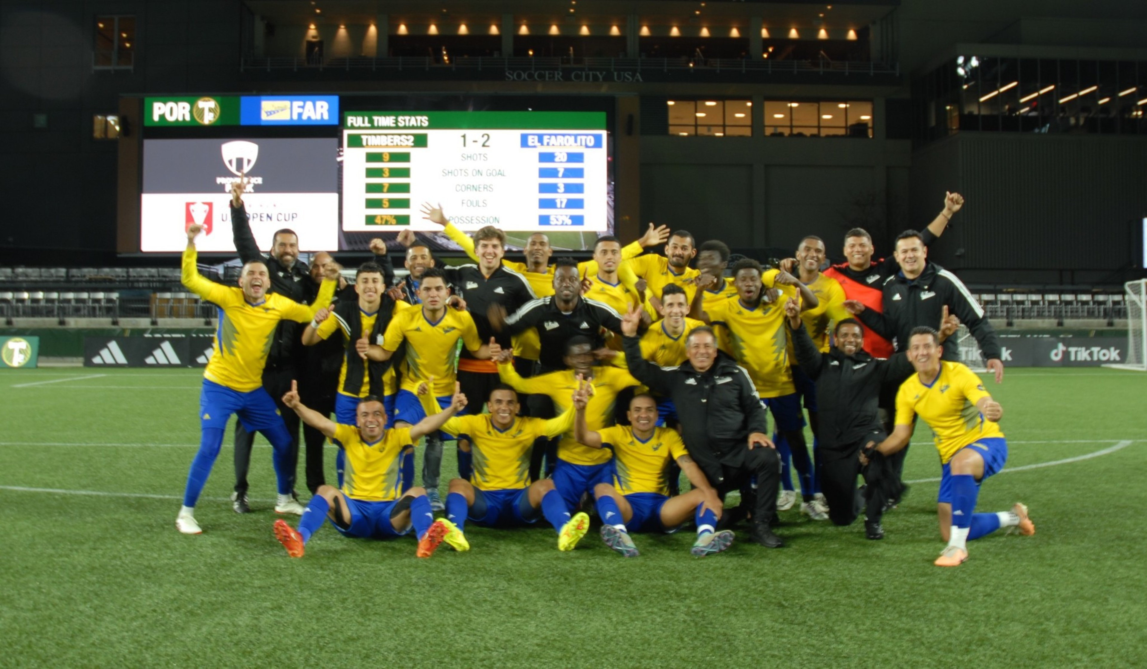 A jubilant soccer team celebrates on a pitch at night with a scoreboard showing a 1-2 victory in the background.