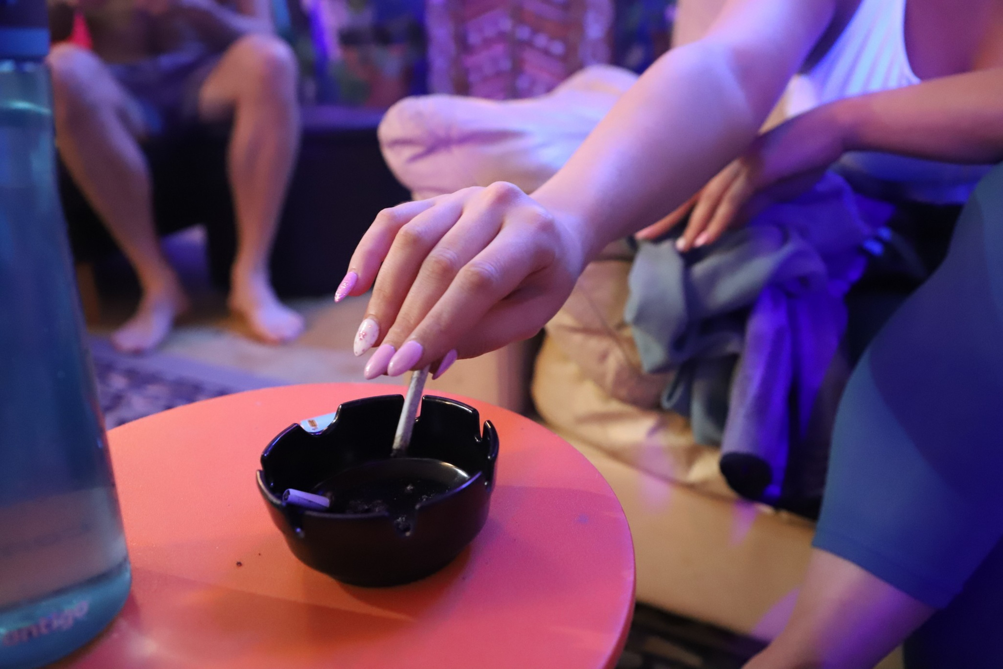 A hand with pink nails extinguishes a cigarette in an ashtray on a red table; background with indistinct seated people.
