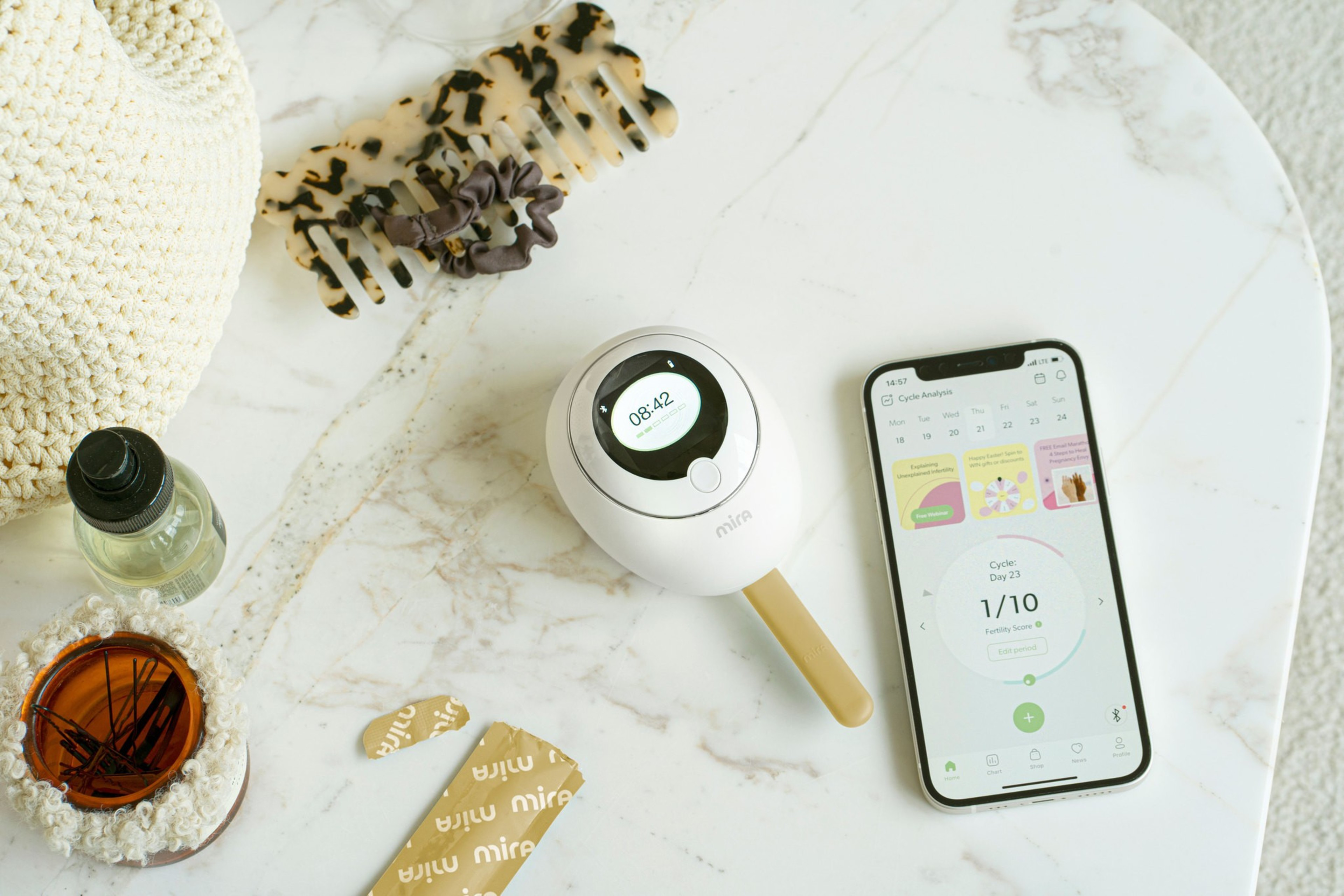 A fertility device and a smartphone with a cycle tracking app sit on a marble surface, surrounded by a knitted bag, a hair clip, an oil bottle, and a plant in a coaster.