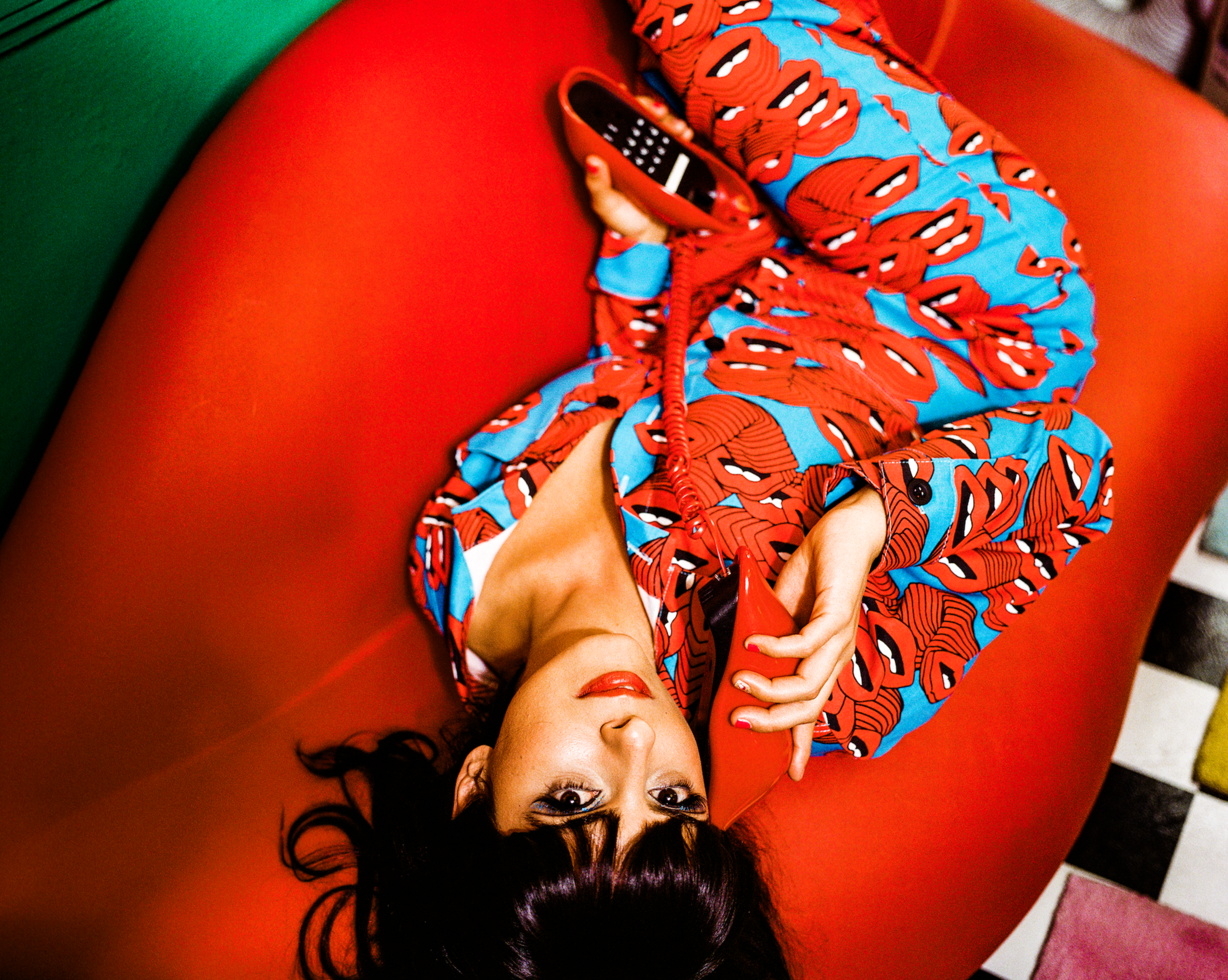 A person lies on a red couch, wearing a blue outfit with red lip prints, holding a red rotary phone handset to their ear.