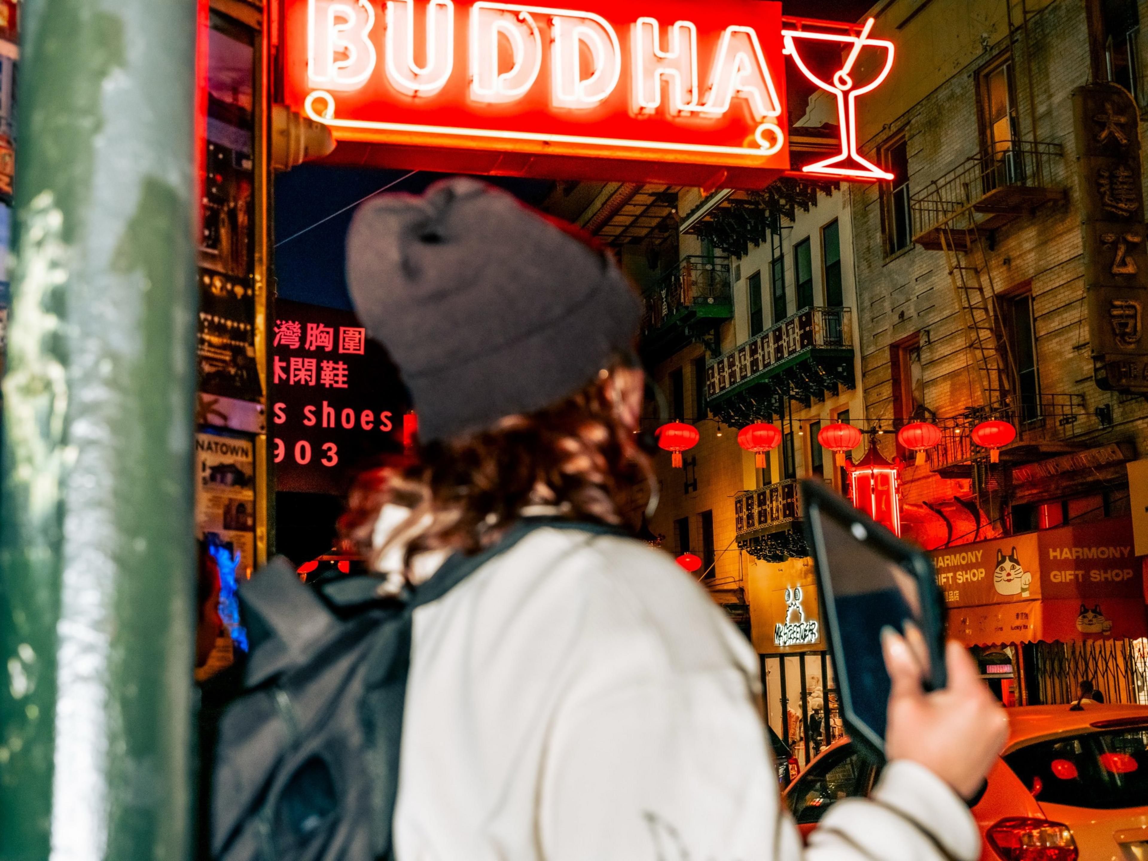 A person with a backpack and holding a tablet stands in front of vibrant neon signs and red lanterns in an urban street, with a &quot;BUDDHA&quot; sign overhead.