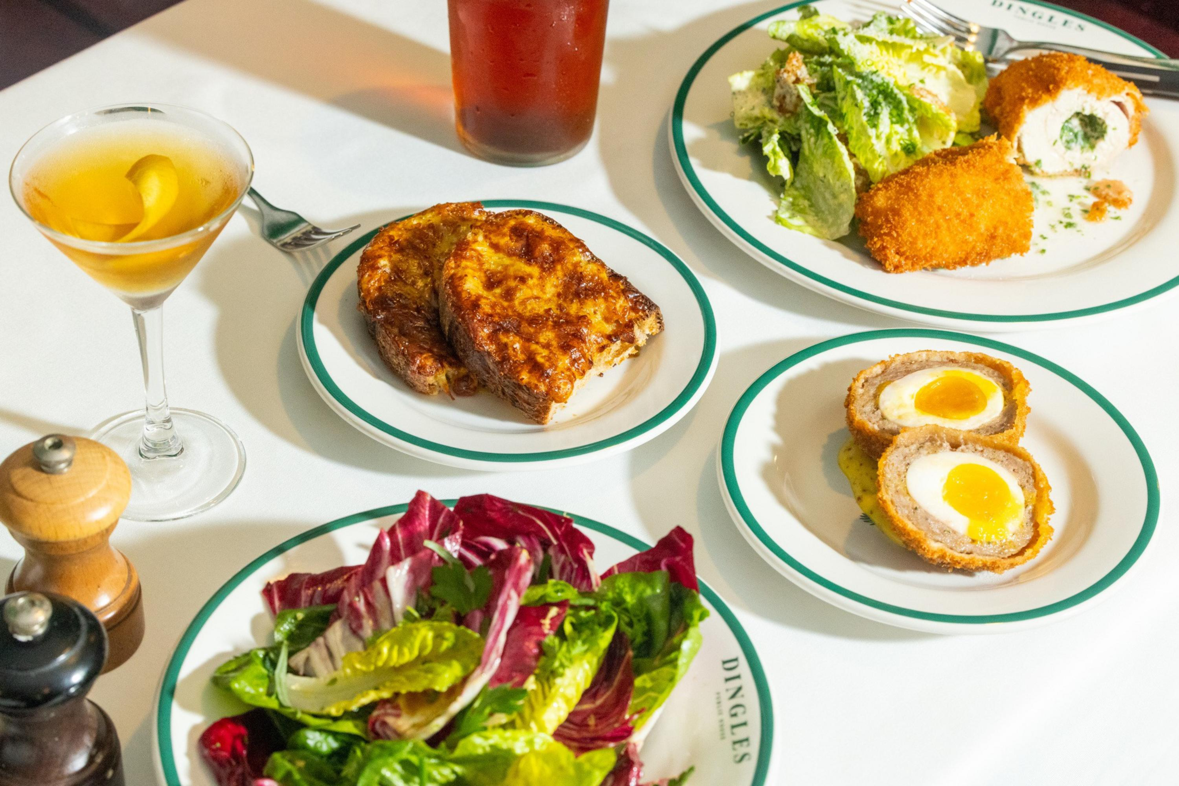Two breaded Scotch eggs, Caesar salad with two breaded rolls, toasted bread slices, mixed green salad, a red drink, and a cocktail with lemon peel on a white table.