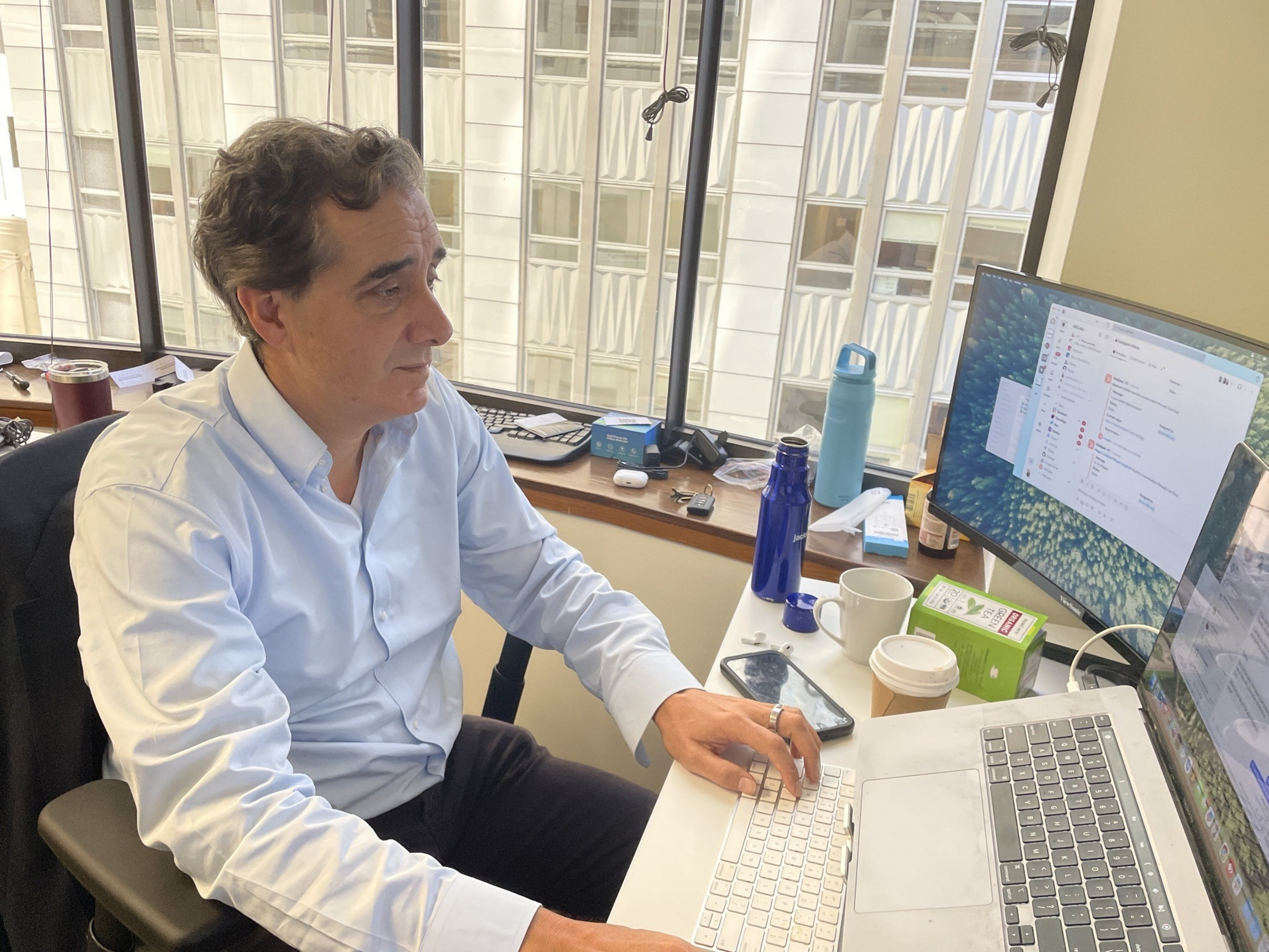 A person in a blue shirt is working at a desk with a laptop, multiple monitors, and various items like bottles and cups. Large windows reveal tall buildings outside.