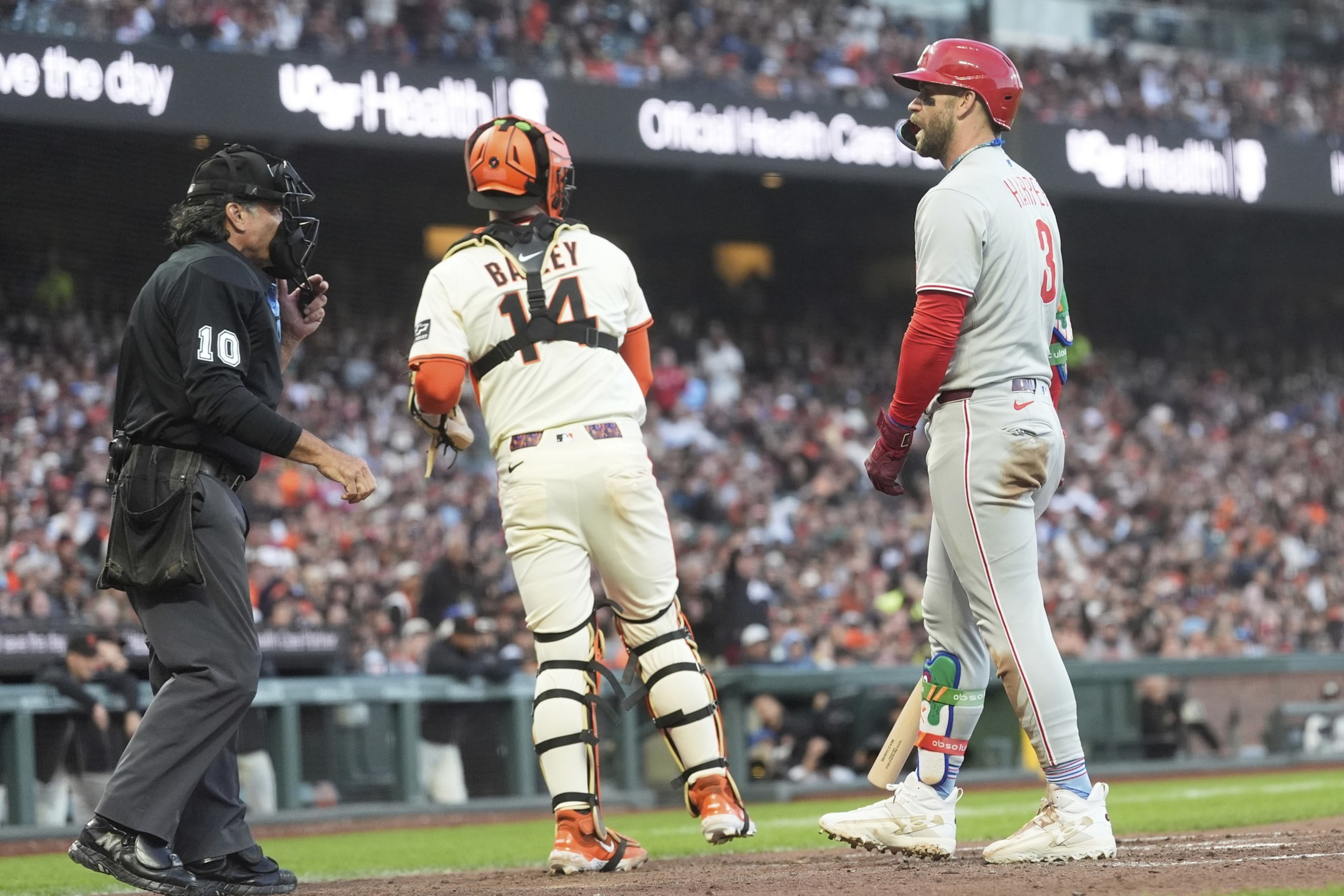 A baseball player in a gray uniform stands at the plate with a bat, facing a catcher in white and orange gear. An umpire in black stands to the side.